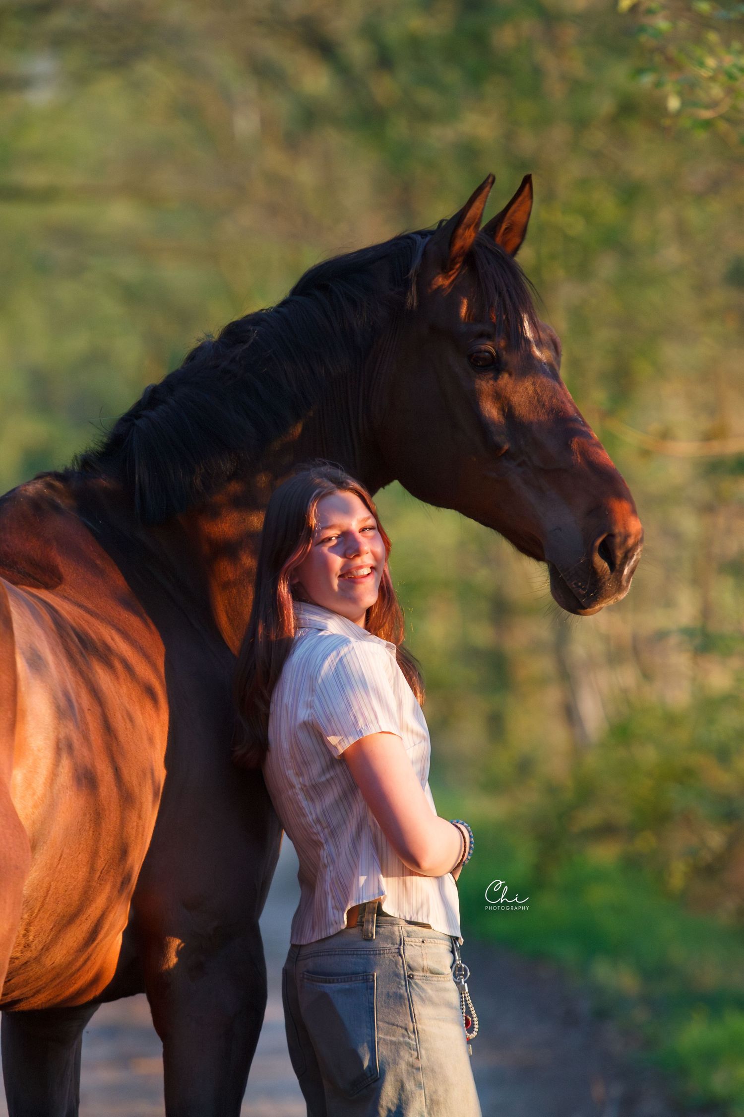 Young woman smiling while standing next to a bay horse on a sunlit path with green trees.