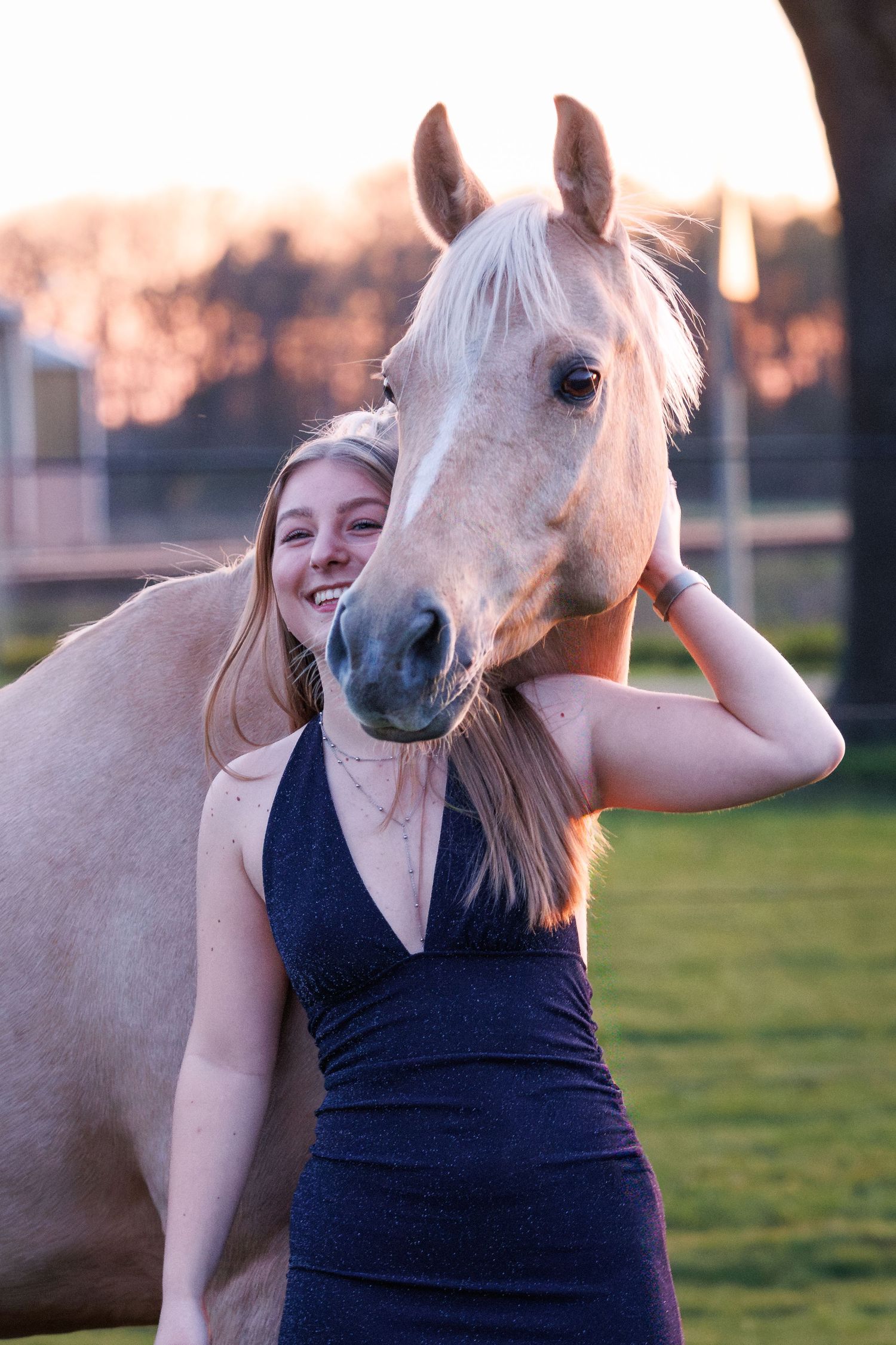 A smiling young blonde woman in a navy dress stands beside a palomino horse at golden hour.