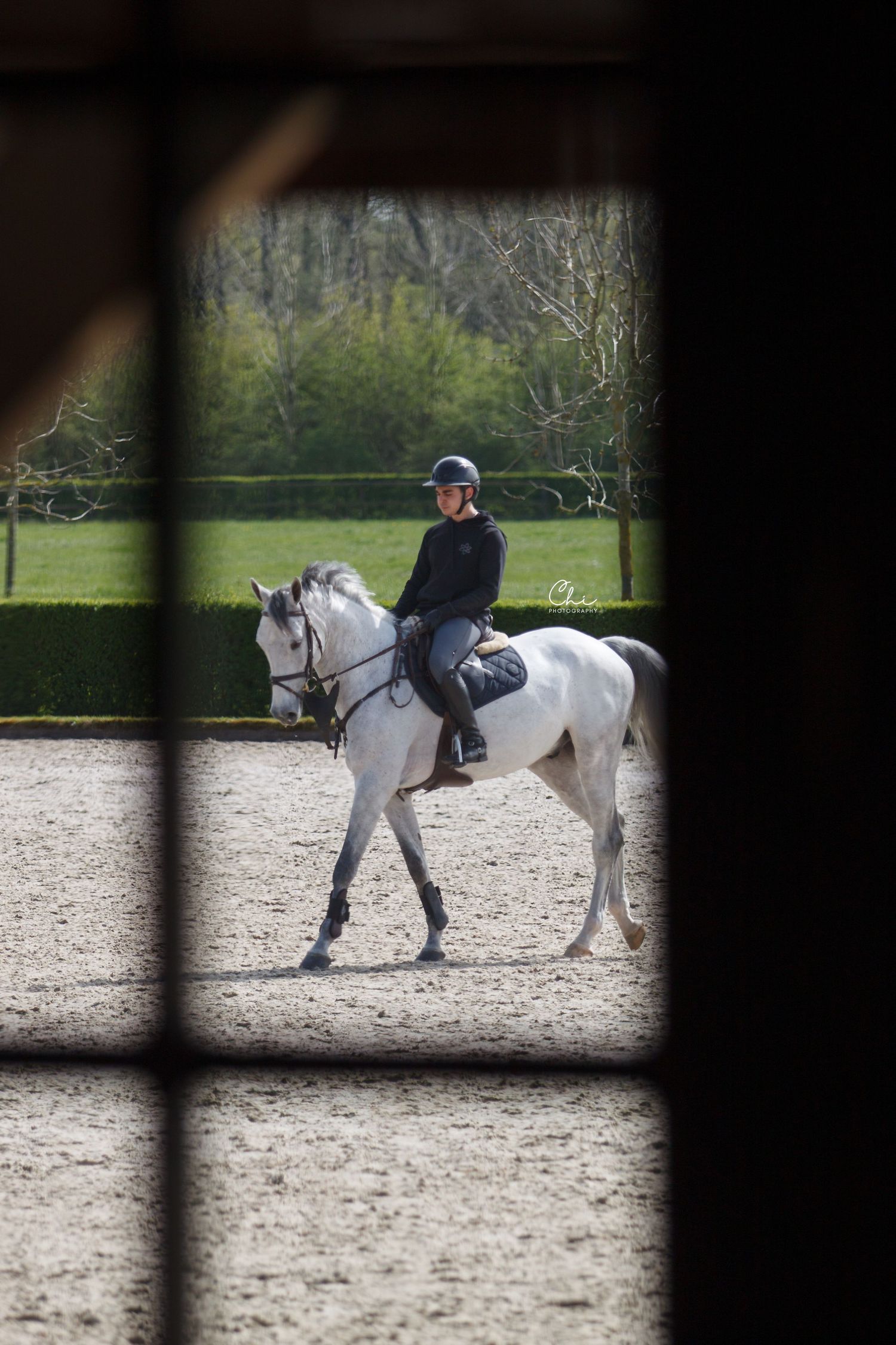 Rider on a grey horse walking in a outdoor arena, viewed through a stable window frame.