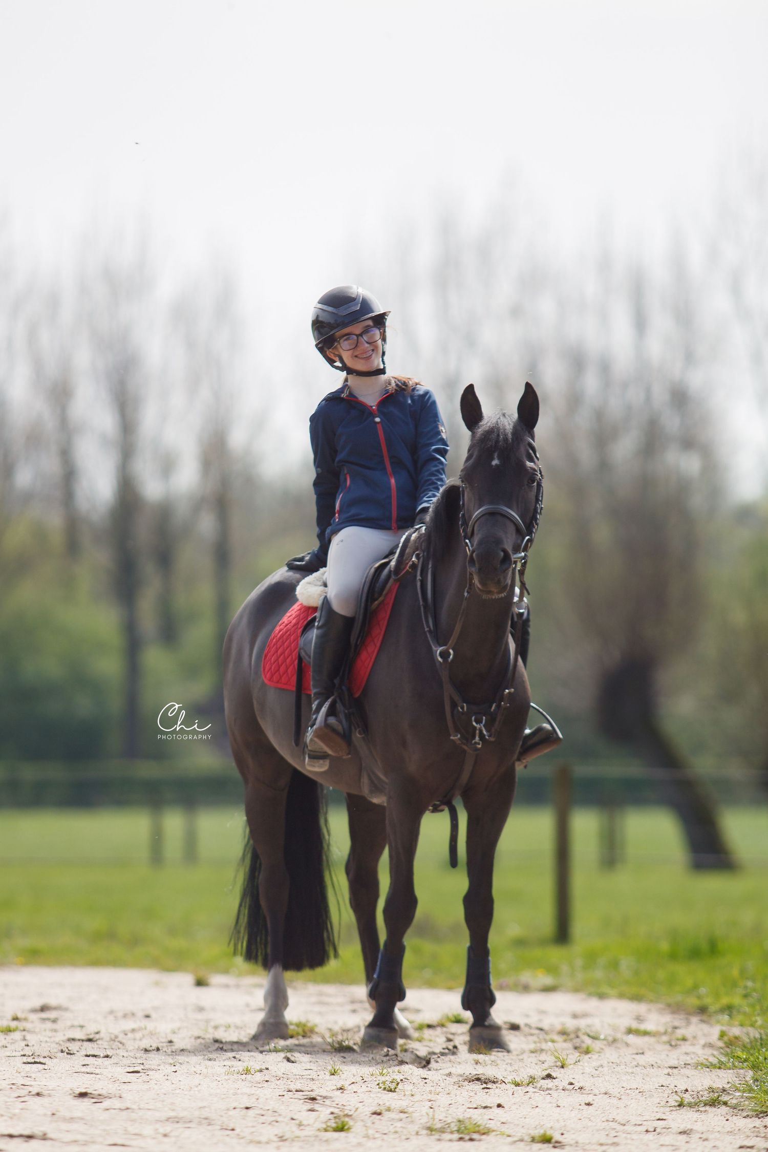 A young female equestrian in a blue jacket rides a dark horse in an outdoor arena surrounded by trees.