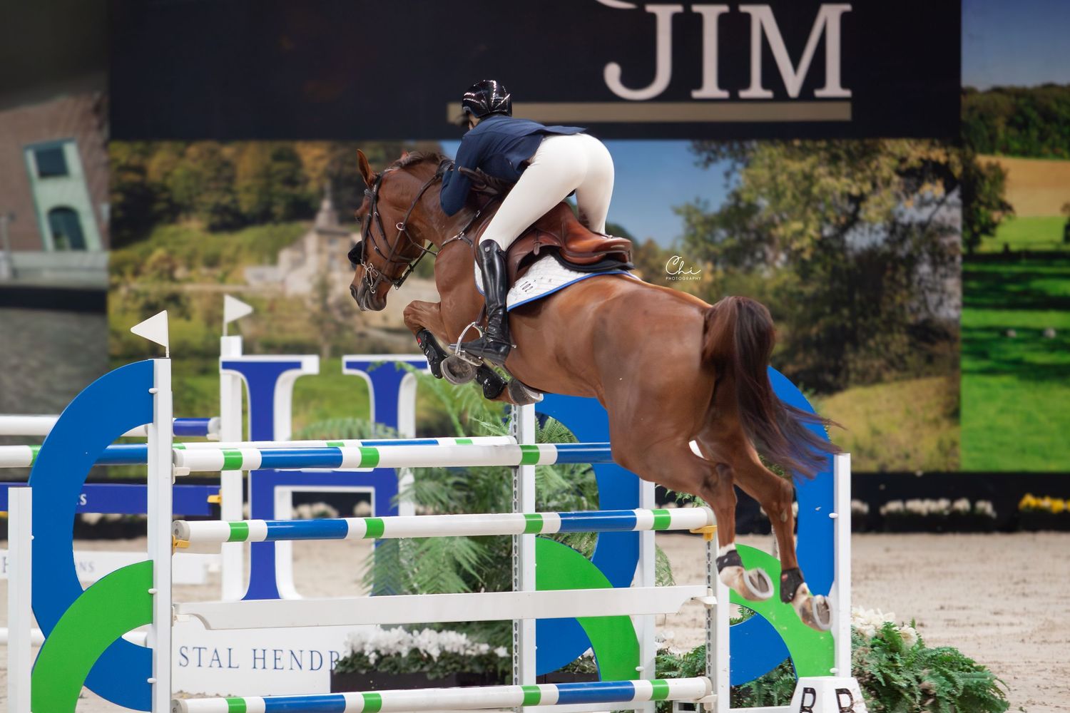 An equestrian competitor on a chestnut horse soars over a colorful jump in an indoor arena competition.
