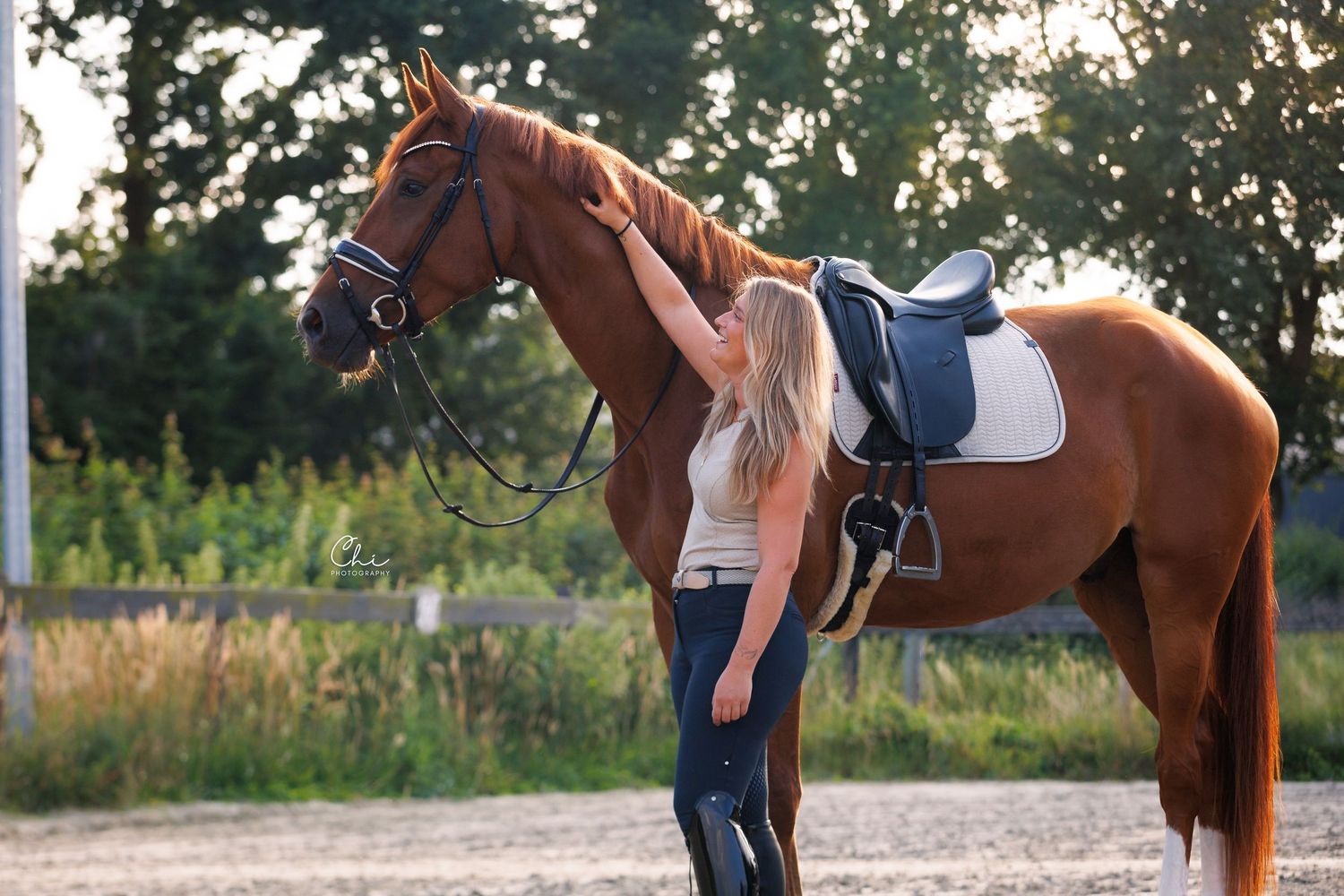 A blonde woman gently strokes a chestnut horse wearing a saddle and bridle in an outdoor riding arena.