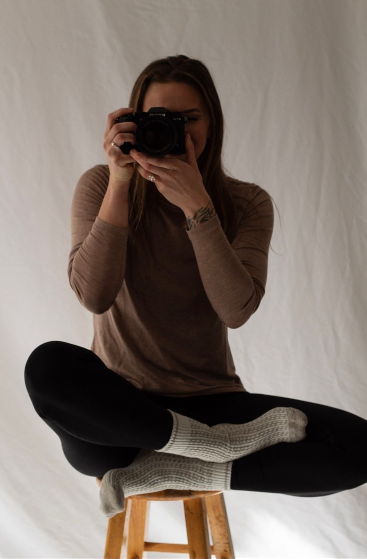 Person in brown sweater and black pants sits on wooden stool taking photos with vintage camera against white backdrop.