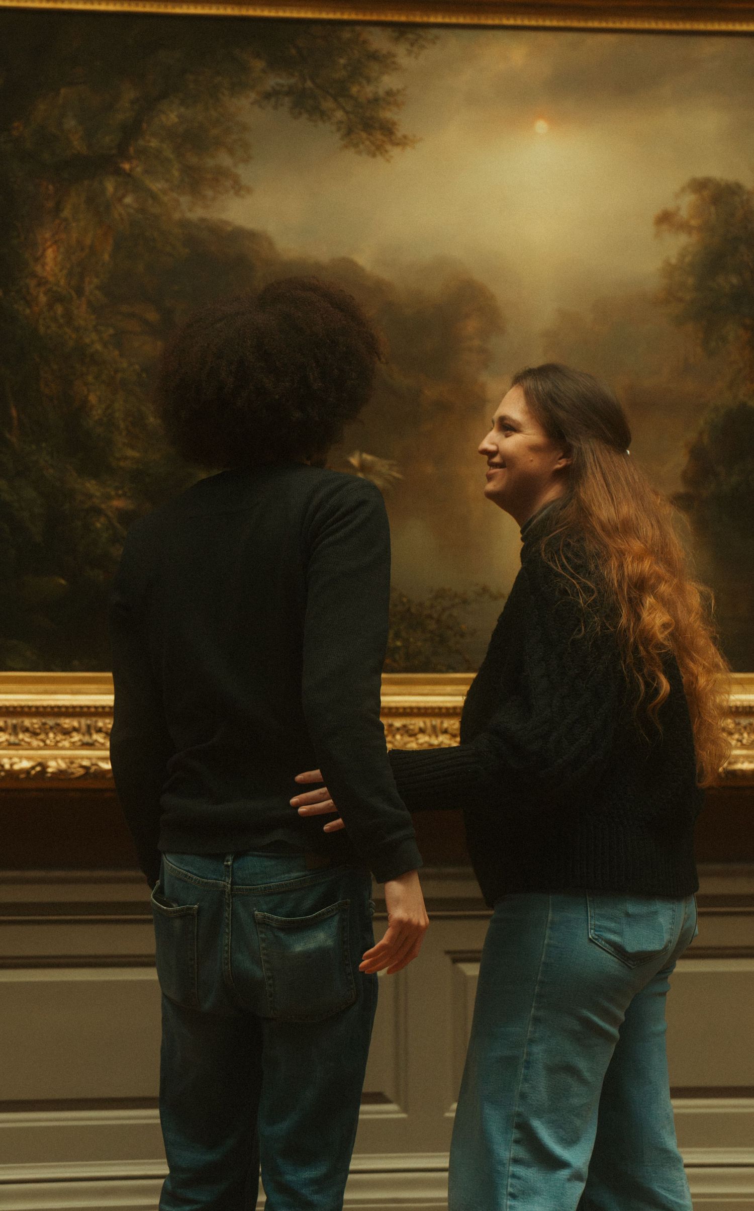 Two people in casual clothing stand together against a dark background with golden lighting in a museum-like setting.
