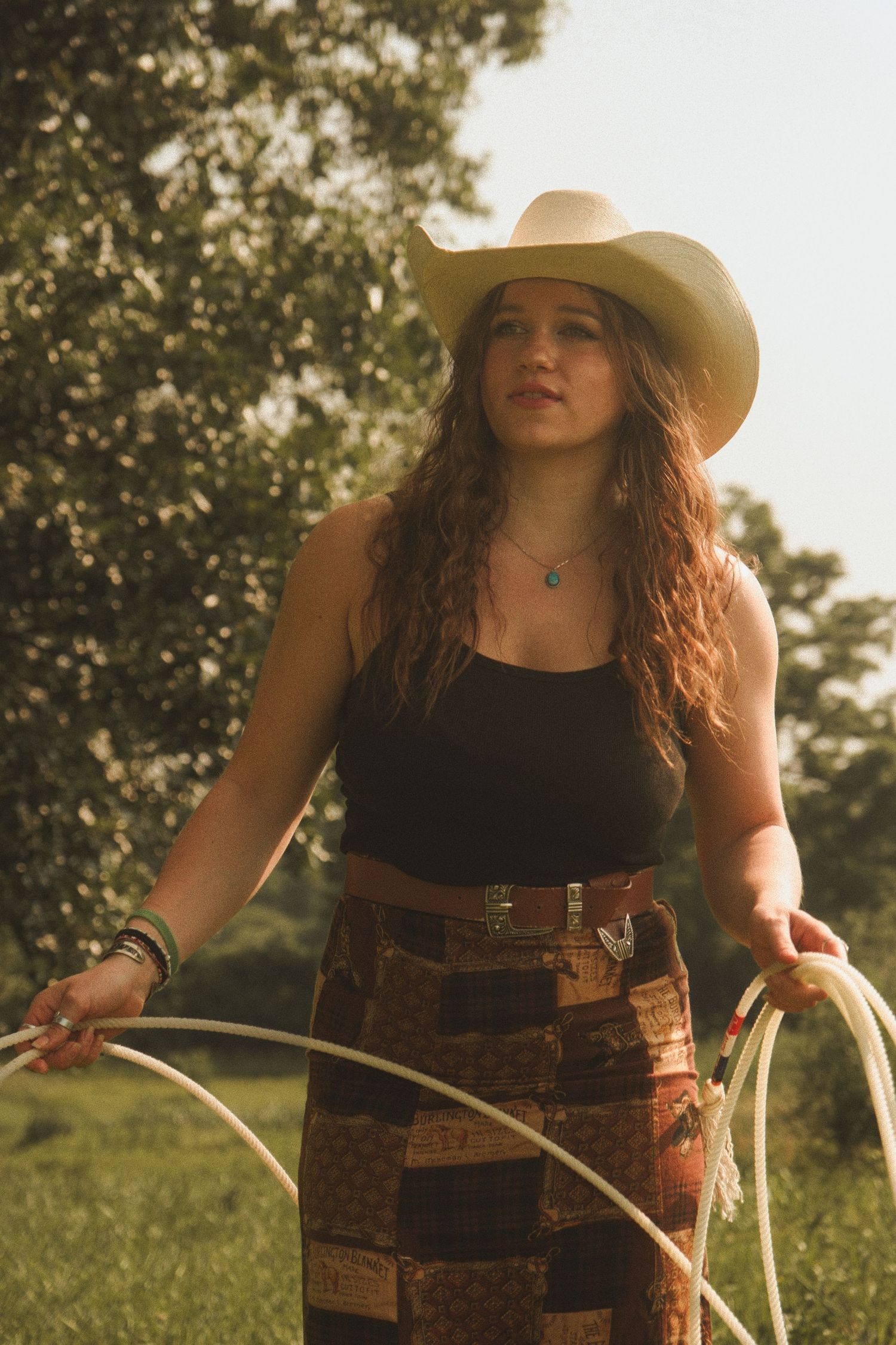 A person in a black tank top and brown leather corset poses outdoors with a bow in golden sunlight.