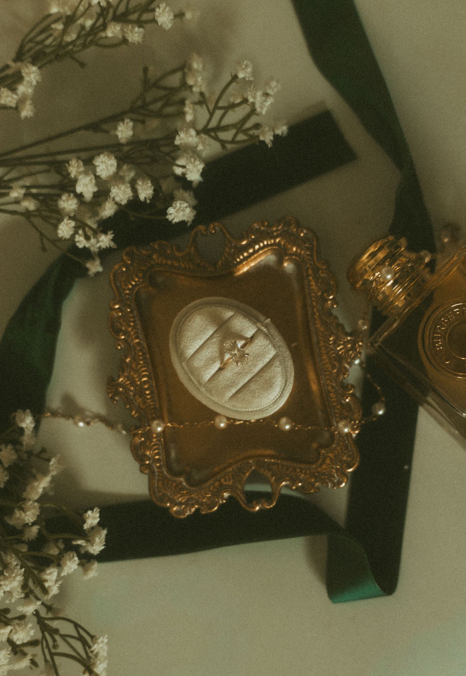Ornate gold vintage ring box with diamond ring, surrounded by baby's breath flowers and dark green ribbon.