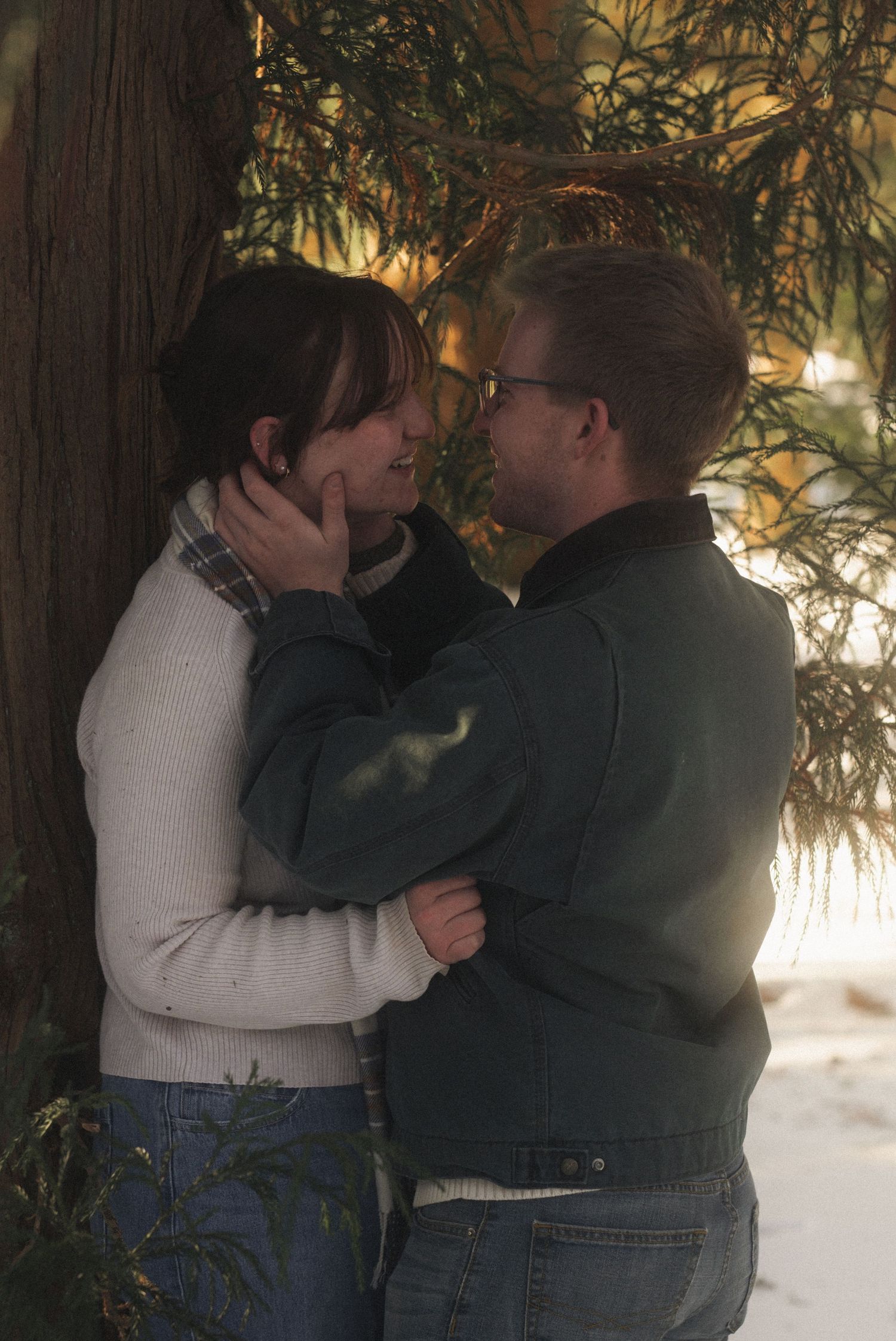 Two people in winter clothing embrace outdoors in a snowy setting with trees in the background.