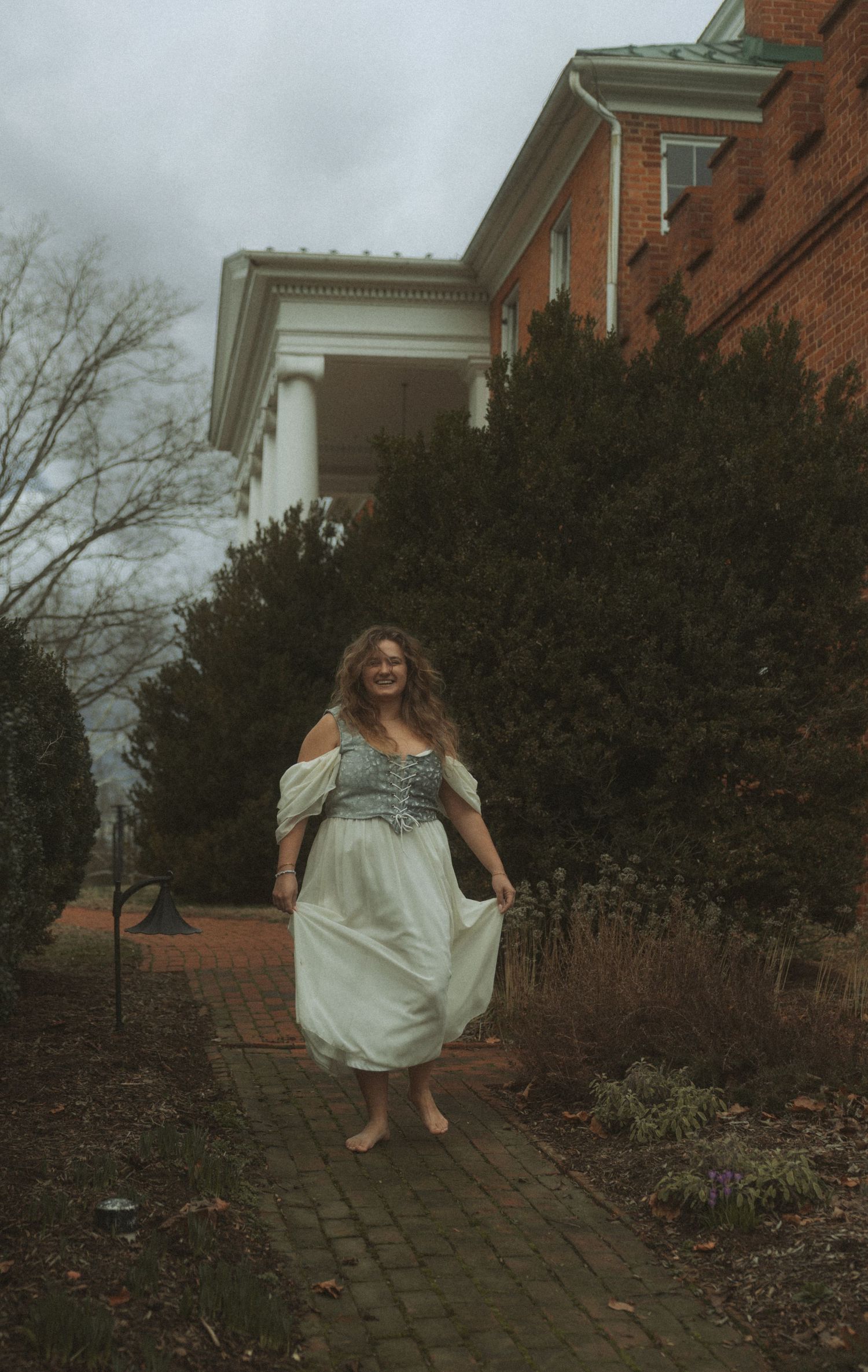 Someone in a flowing white dress twirls gracefully on a brick pathway surrounded by winter garden foliage.