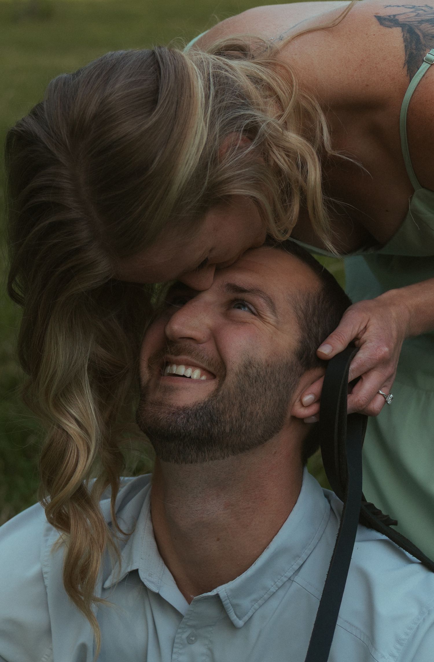 A smiling bearded man looks up as a blonde woman leans down to kiss his forehead outdoors.
