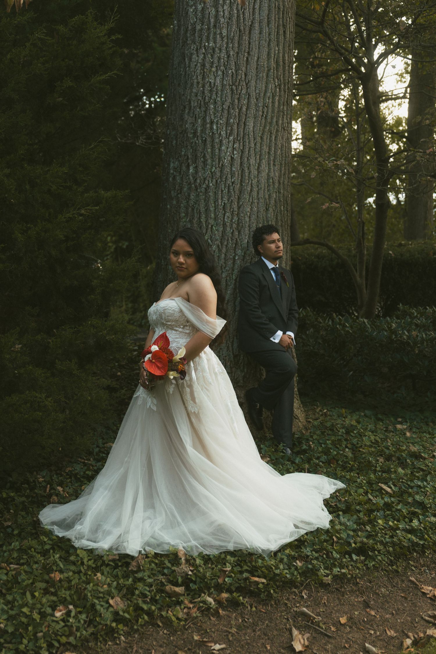 Bride in white off-shoulder gown holding red bouquet stands before groom in dark suit, posing in lush forest setting.