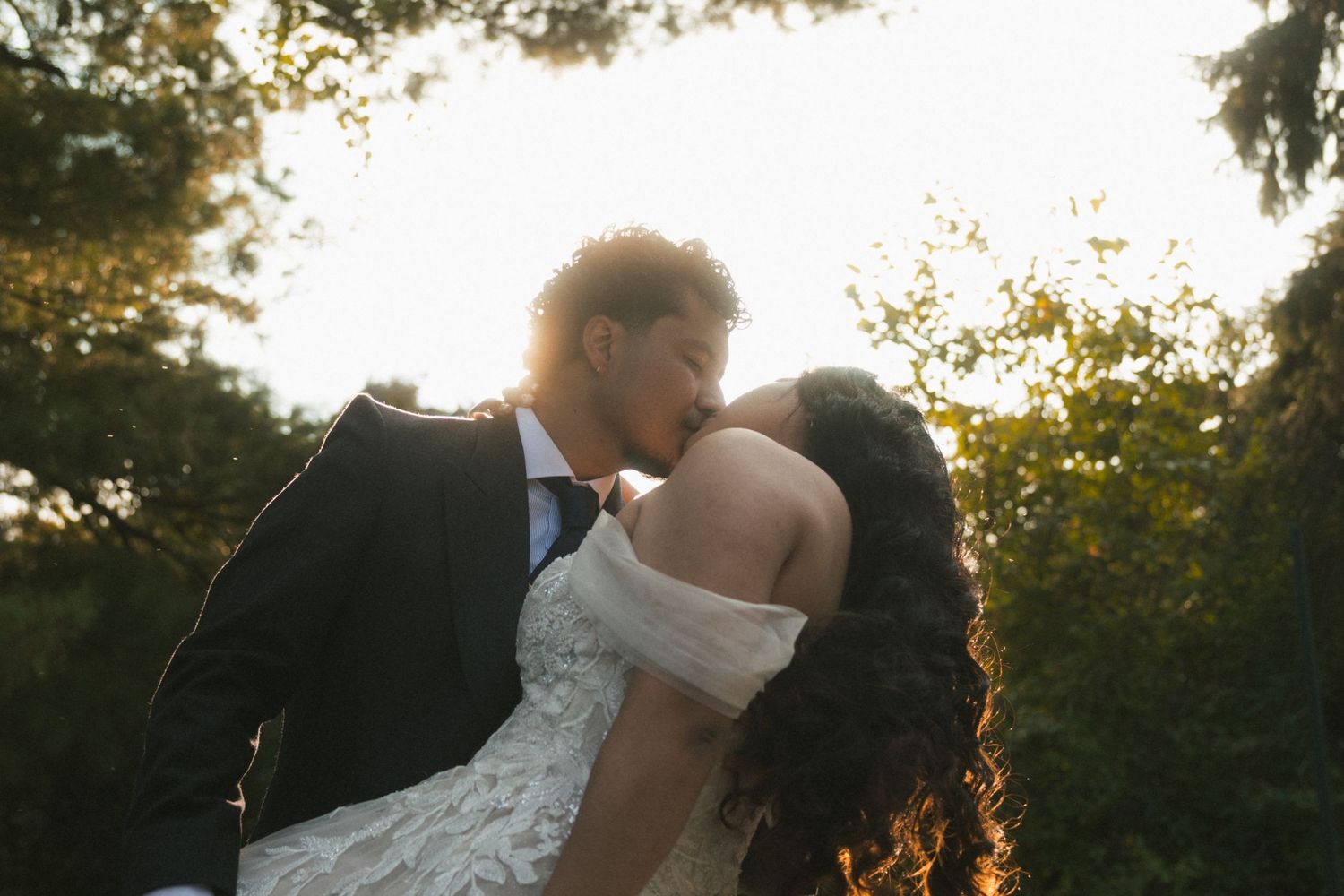 A couple shares a romantic kiss in wedding attire, backlit by golden sunset light with lush green trees behind them.