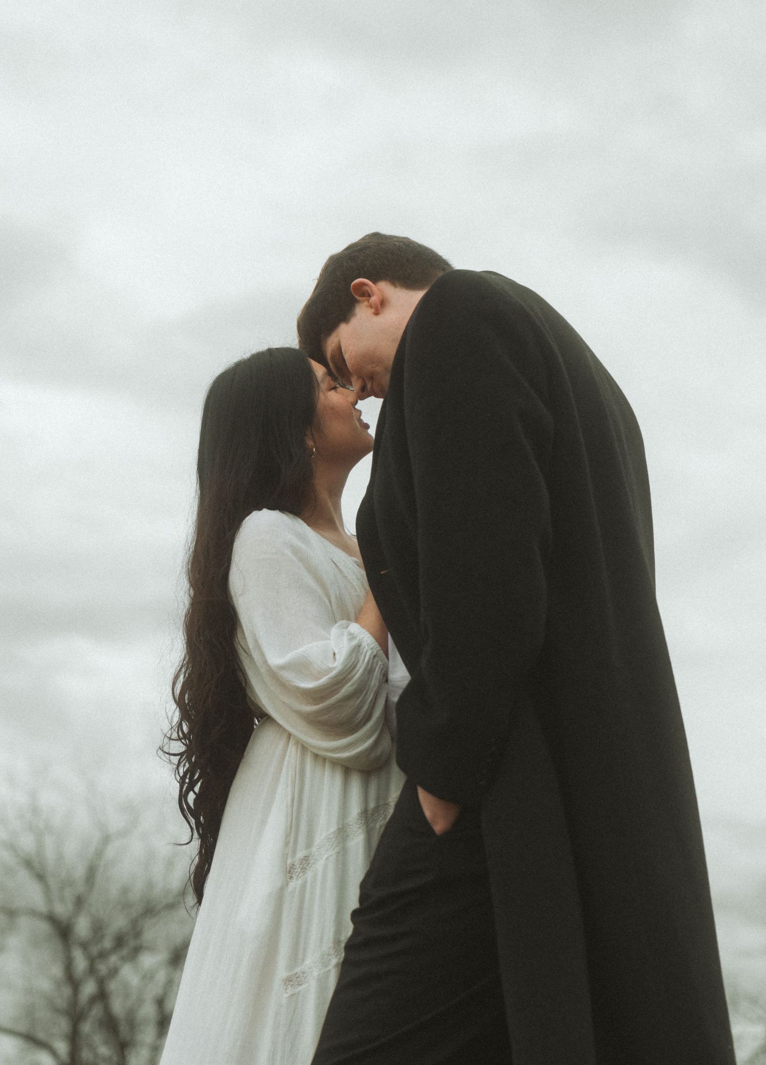A romantic couple in contrasting black and white outfits share an intimate moment against a moody cloudy sky.