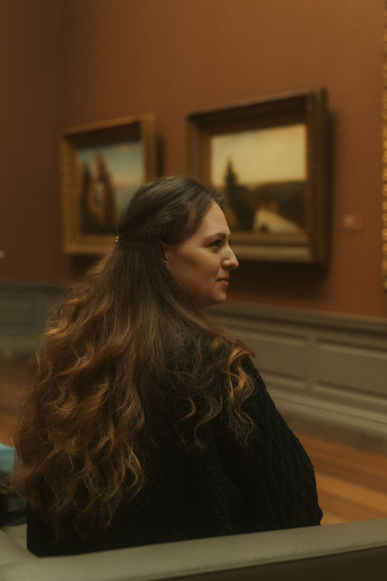 A person with long wavy brown hair sits in an art gallery, viewing paintings on a warm-toned wall.