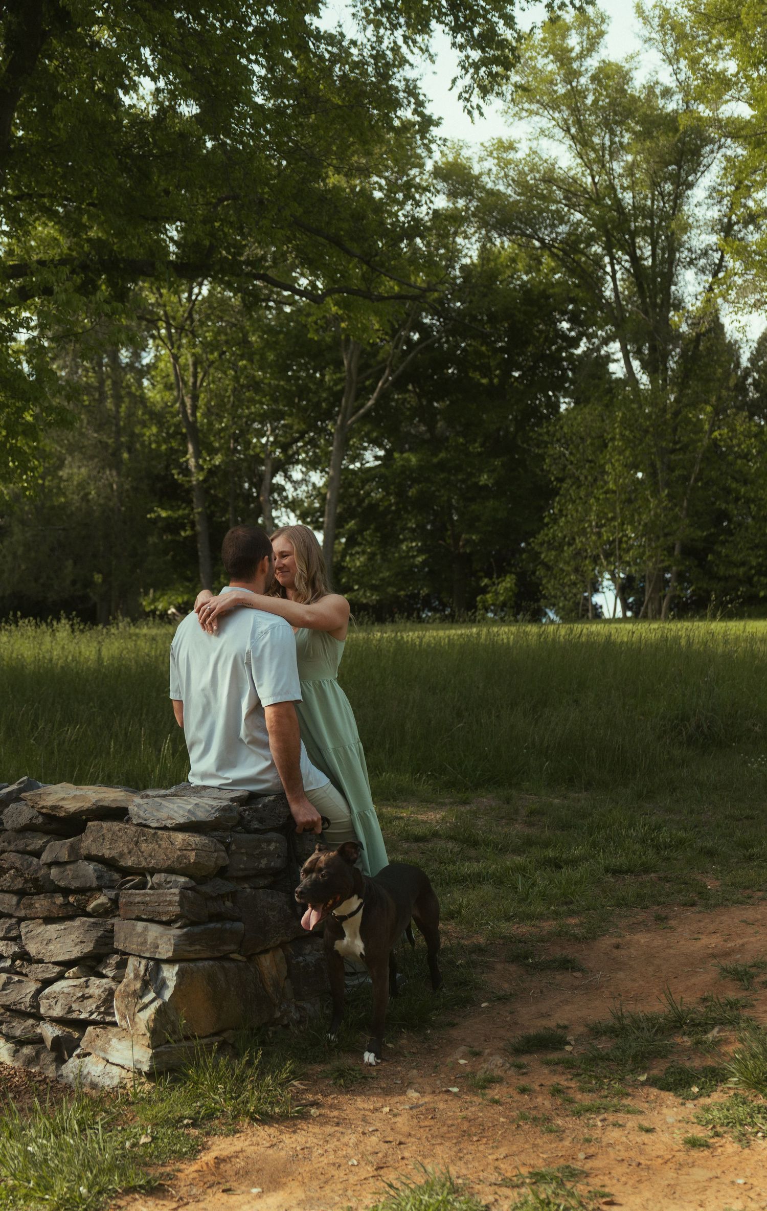 Couple embracing near a stone wall with a dog in a lush green park setting on a sunny day.