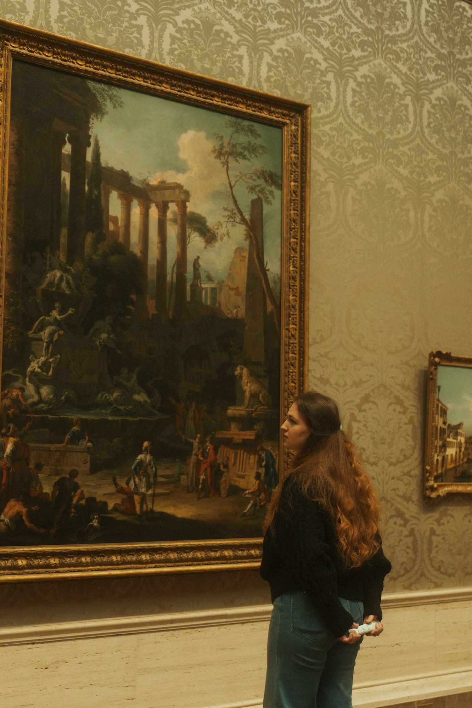 A museum visitor admires a large classical painting of ancient ruins displayed on an ornate wallpapered wall.