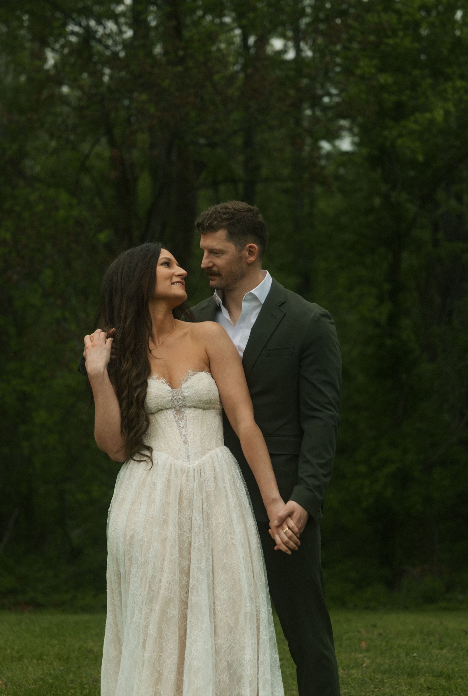 A couple in wedding attire holds hands in a lush green forest, the bride in a white gown smiling as the groom gazes at her.