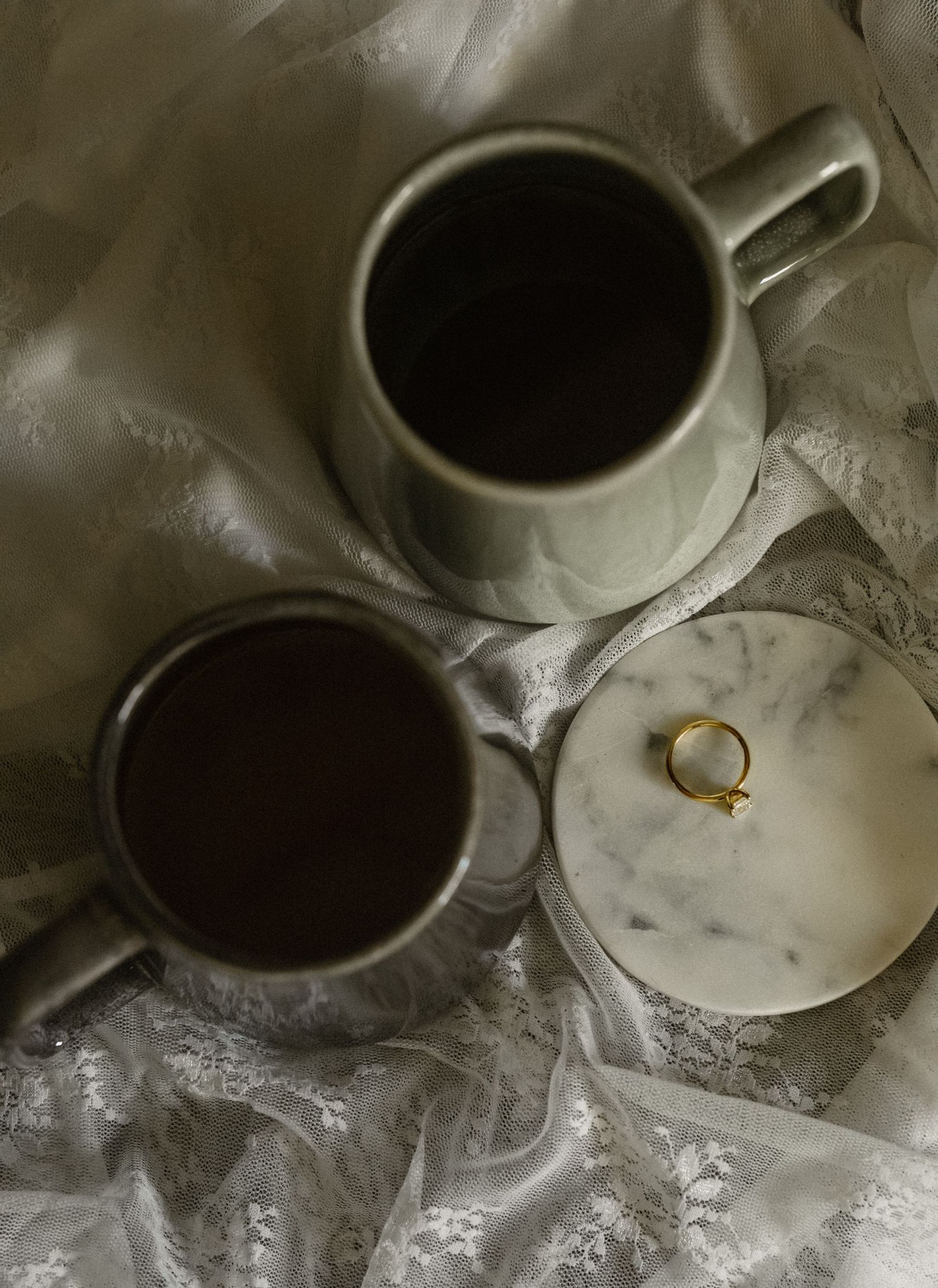 Two cups of black coffee and a gold ring on white fabric with moody lighting and dark shadows.