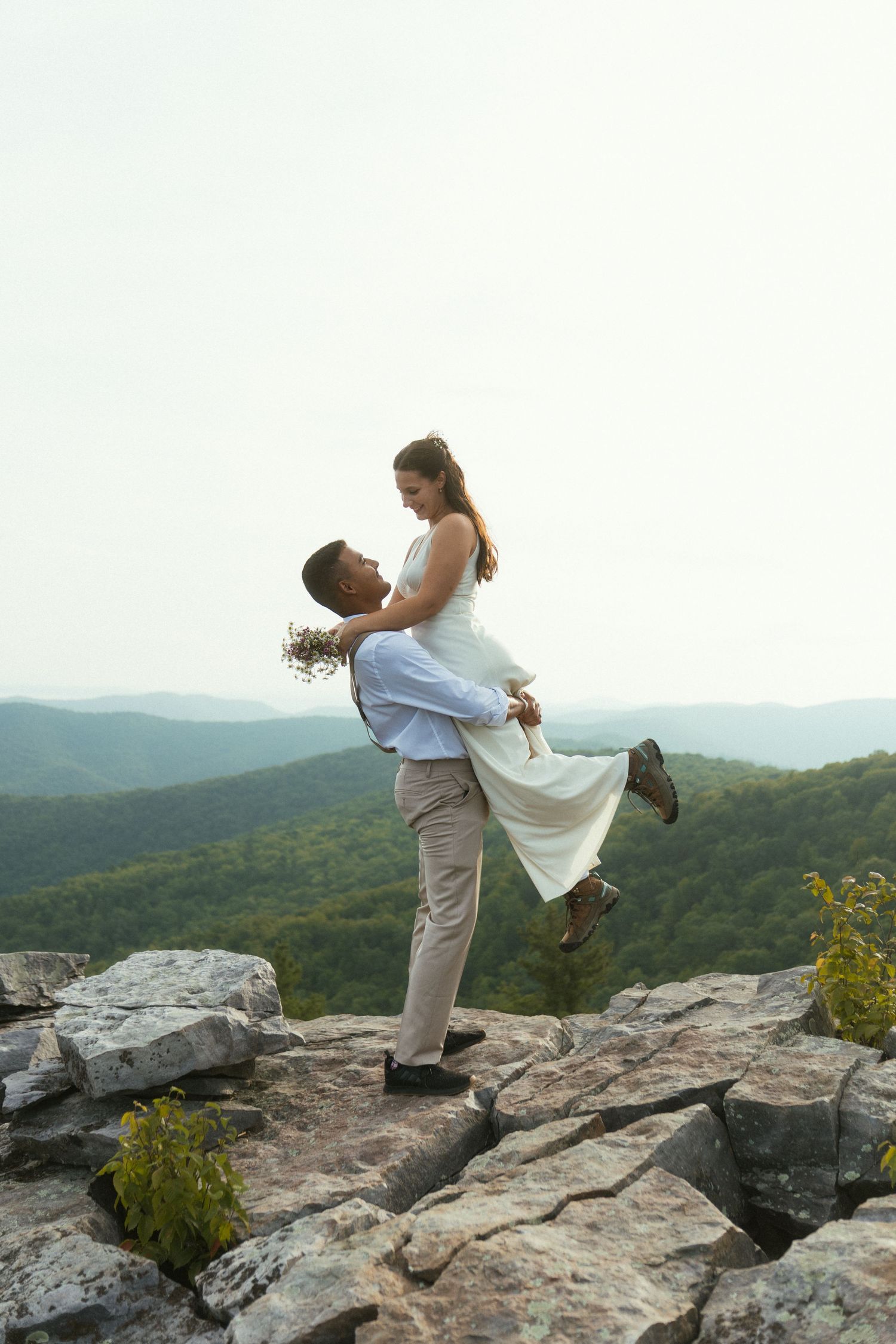 Groom lifting bride on rocky mountain overlook with lush green valley views during outdoor wedding ceremony.