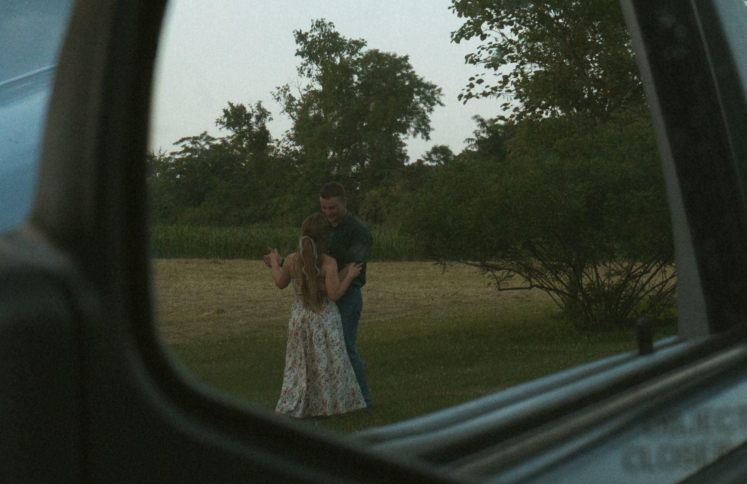 Couple embracing outdoors reflected in a car side mirror, surrounded by lush green trees and grass at dusk.