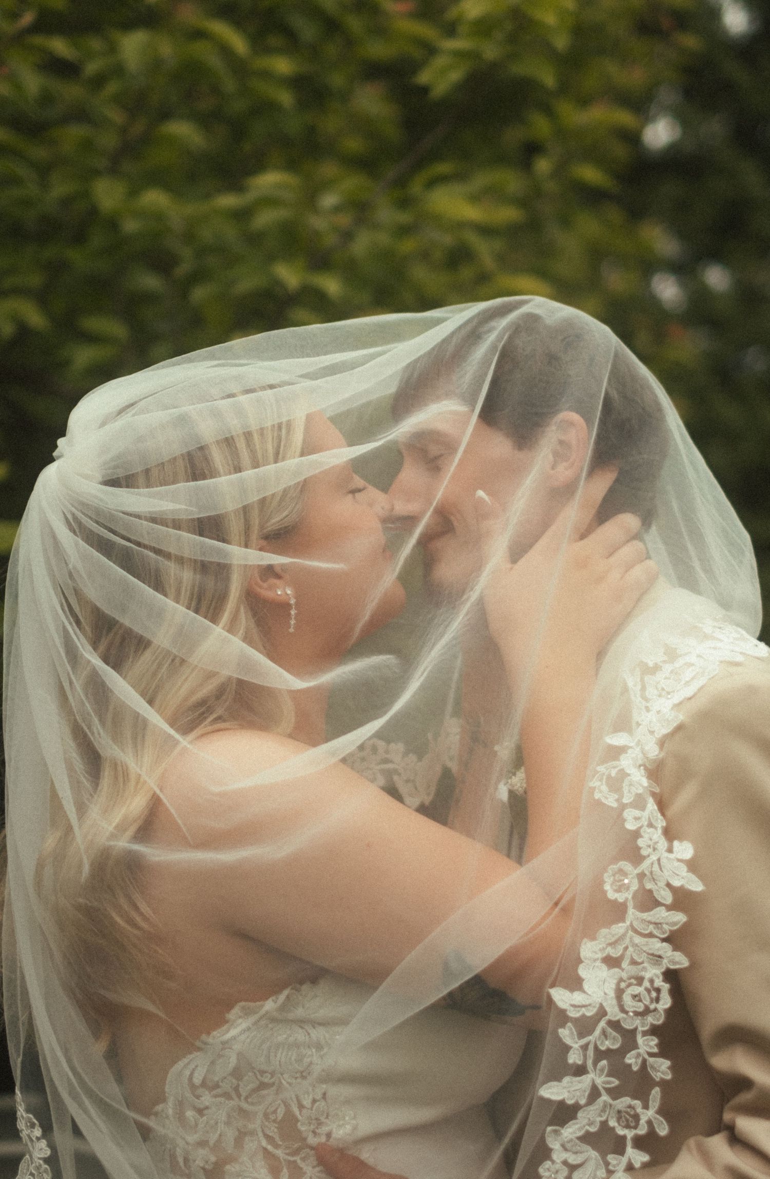 Bride and groom sharing a romantic kiss under a lace-trimmed wedding veil outdoors.