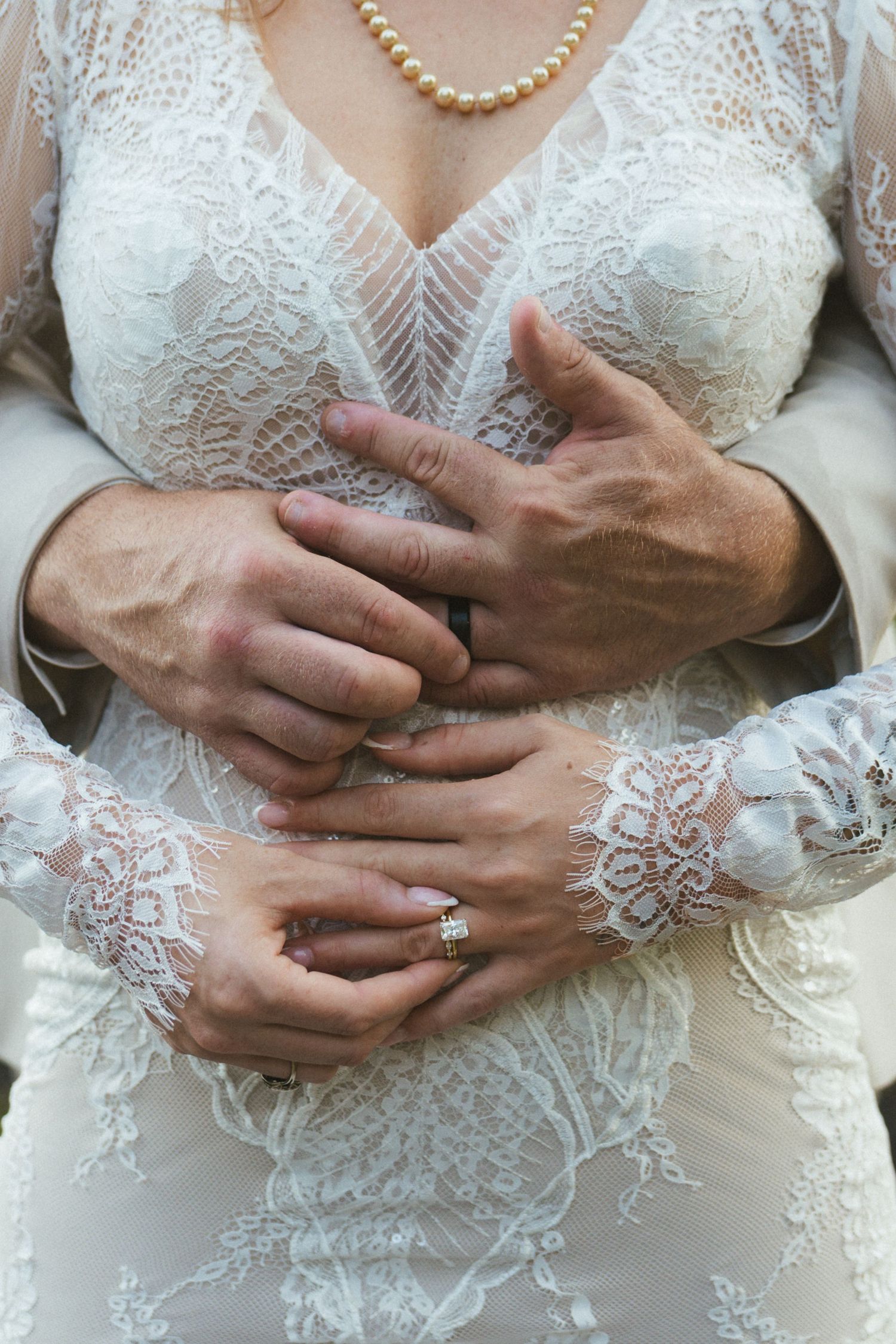 Close-up of bride and groom hands intertwined, bride wearing diamond ring and lace wedding dress with pearl necklace.