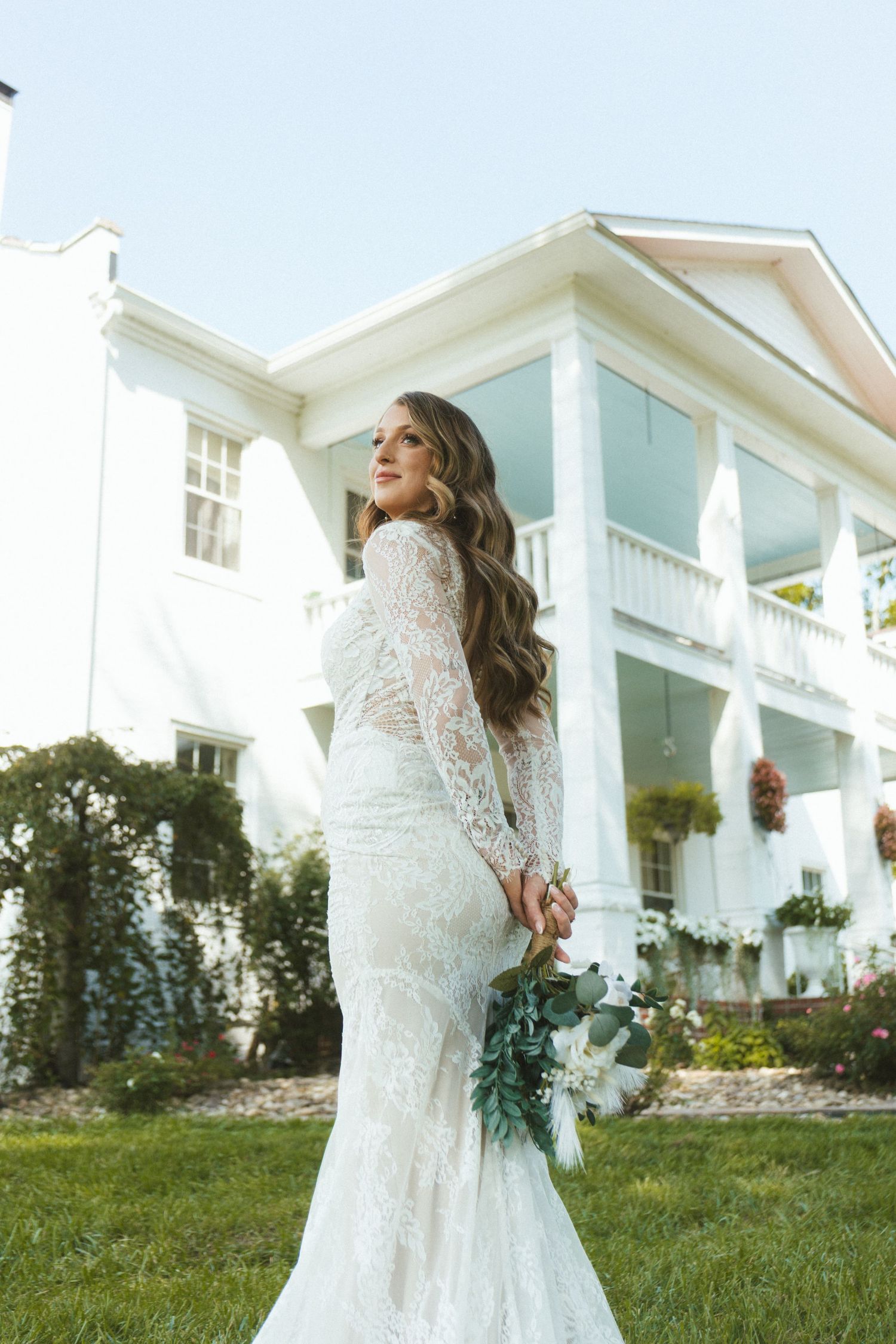 Bride in lace long-sleeve wedding gown holding bouquet, posing in front of elegant white mansion.