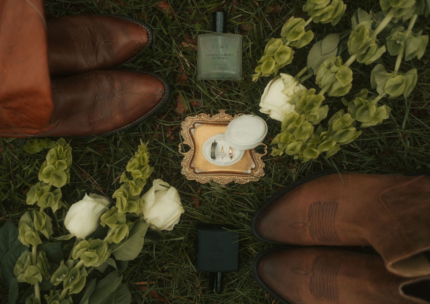 Overhead view of wedding rings in ornate box surrounded by white roses and greenery between two pairs of brown leather shoes.