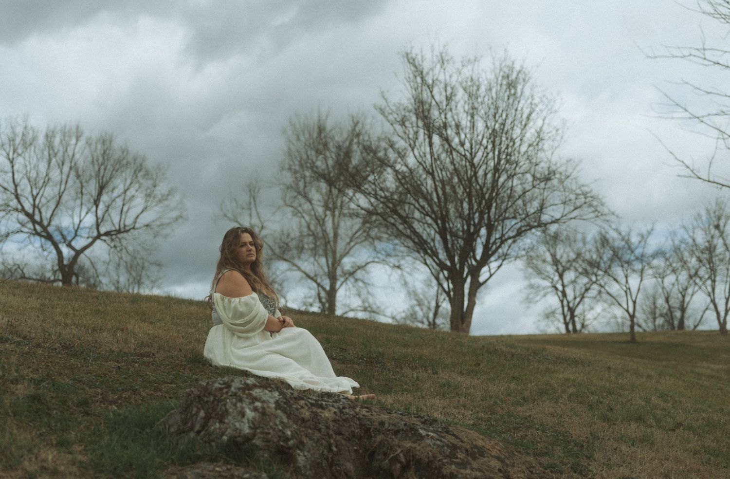 A person in a flowing white dress poses gracefully on a grassy hillside on an overcast day.