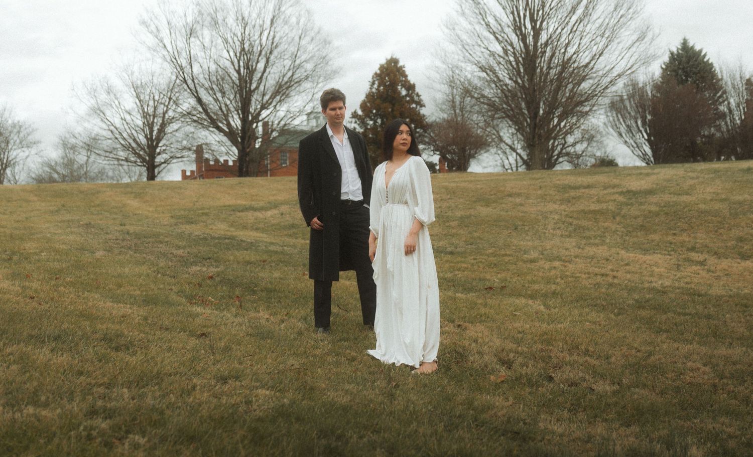 A couple in wedding attire walks together across a grassy hillside on an overcast autumn day.