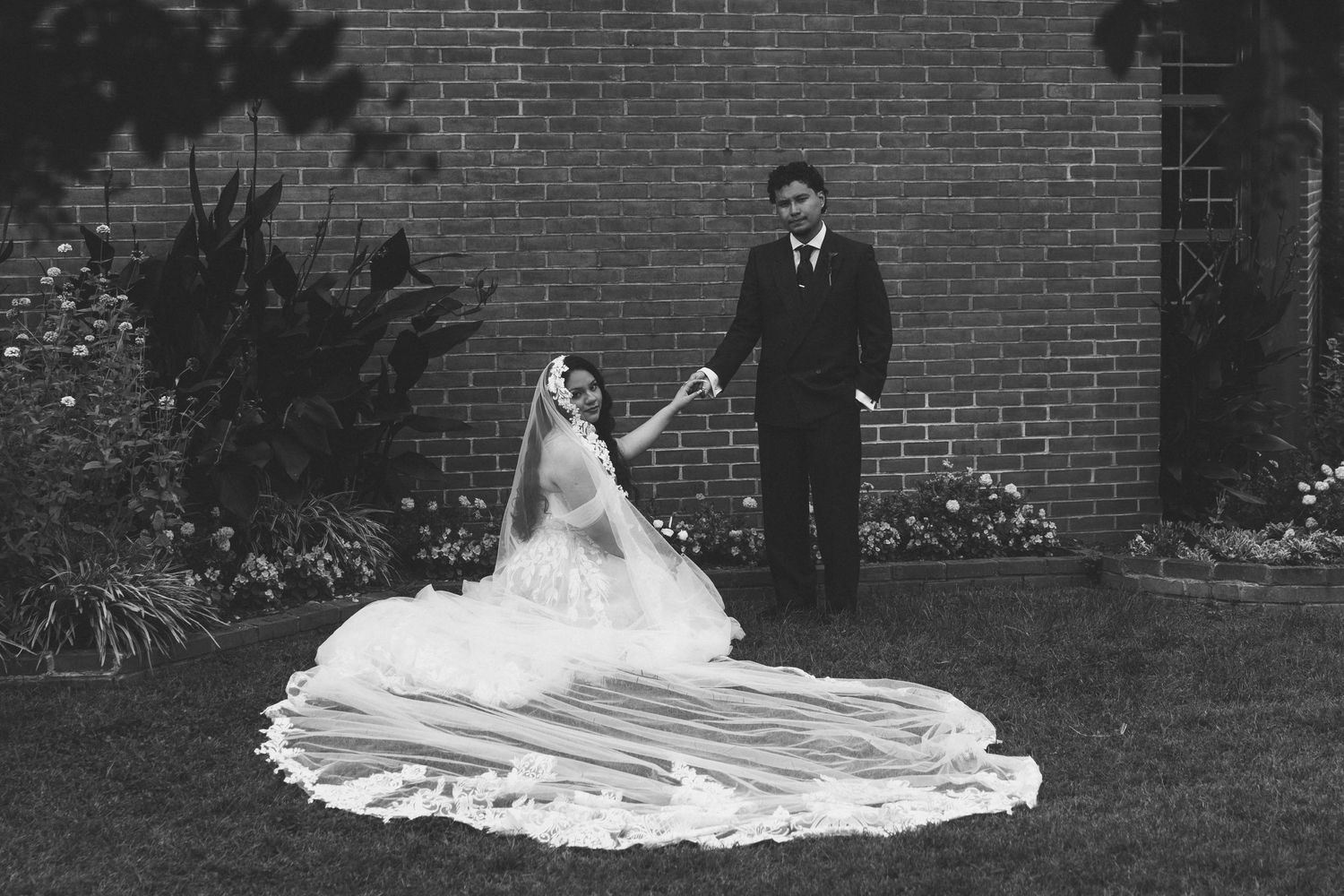 Black and white wedding photo of bride in flowing gown and veil holding groom's hand near brick wall.