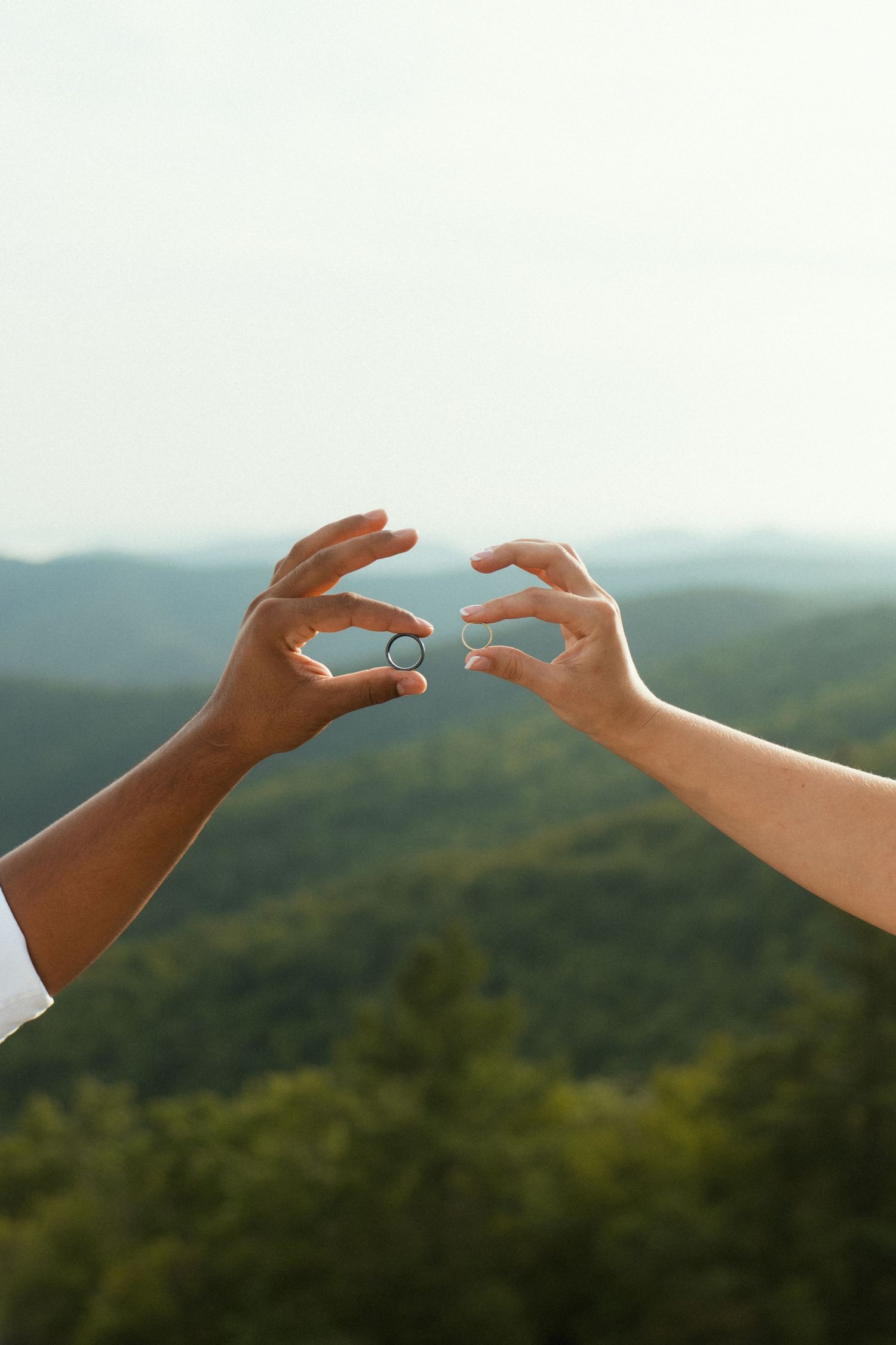 Two hands holding wedding rings against a scenic mountain backdrop with lush green forests below.