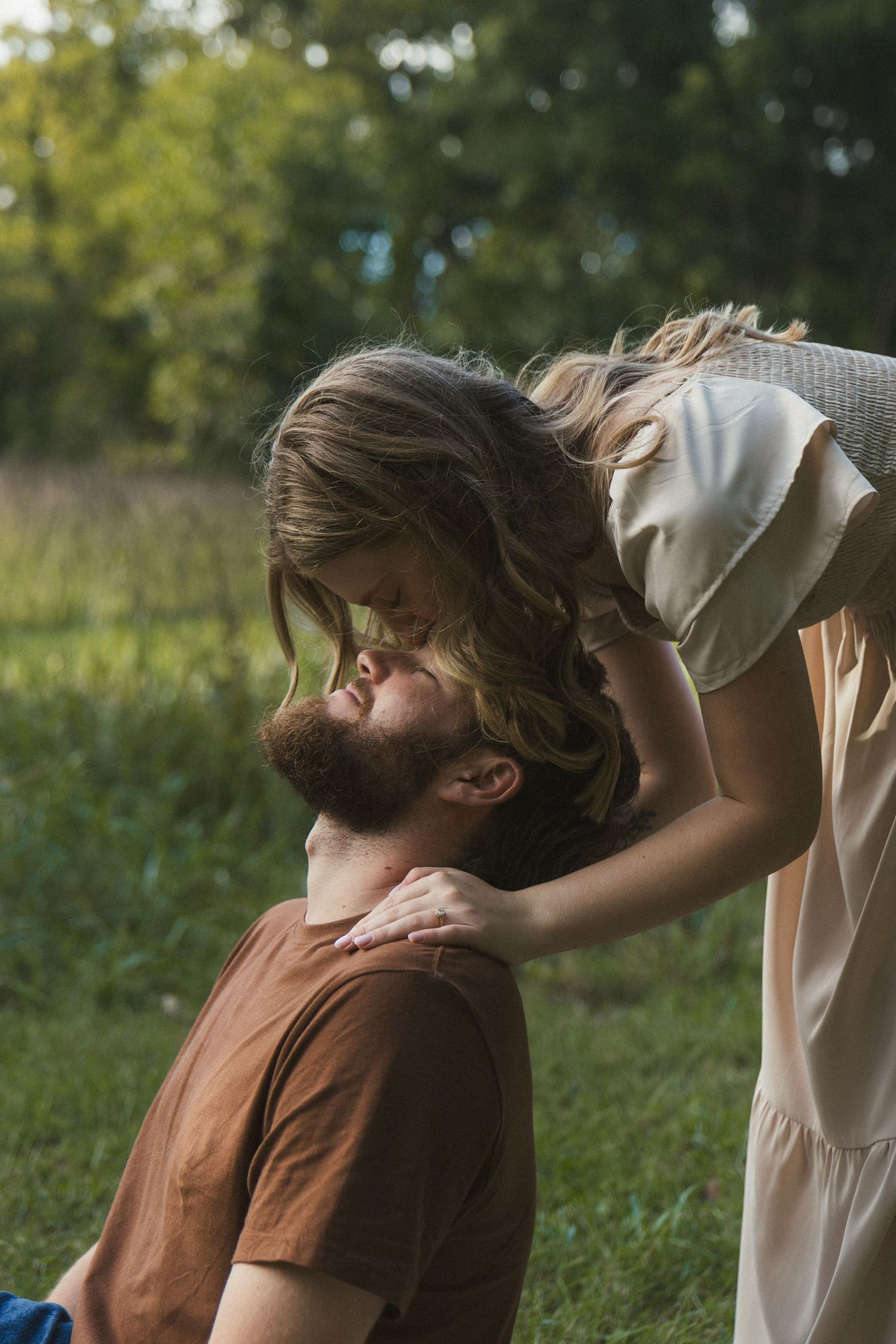 A woman in a white dress leans down to kiss a bearded man in a brown shirt sitting in a lush green field.