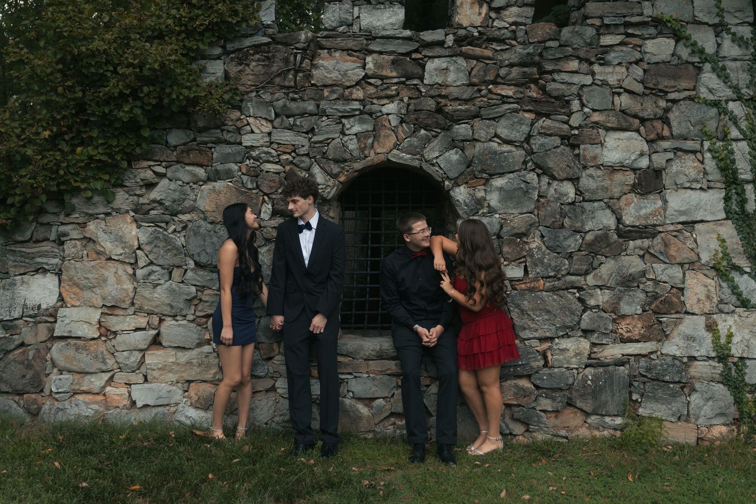 Two couples in formal attire pose before a rustic stone wall with an arched window outdoors.