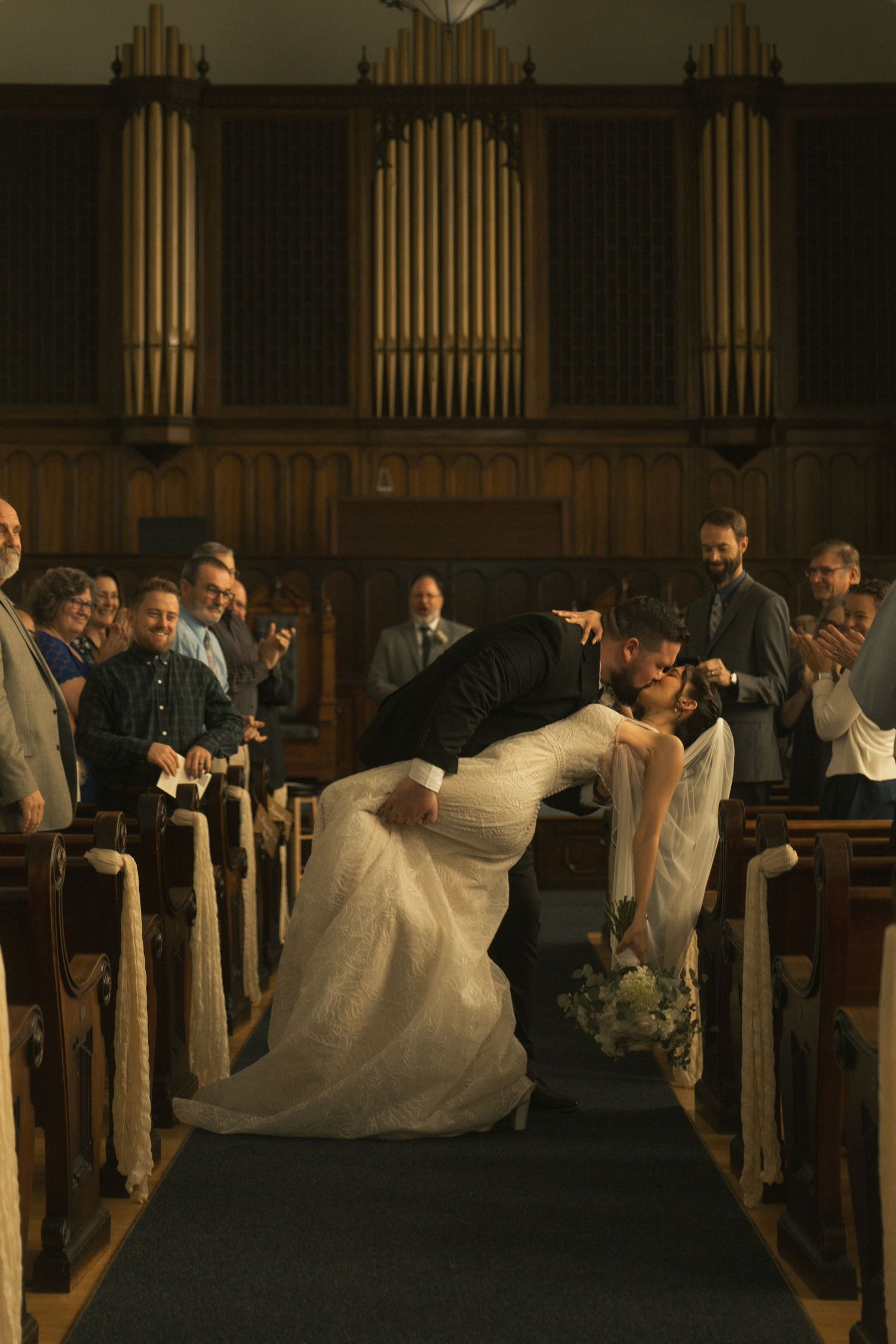 Bride and groom share a romantic dip kiss in church aisle surrounded by applauding wedding guests with pipe organ backdrop.