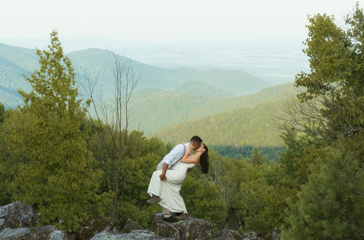 Couple sharing a romantic kiss on a rocky mountain overlook surrounded by lush green trees and scenic valley views.