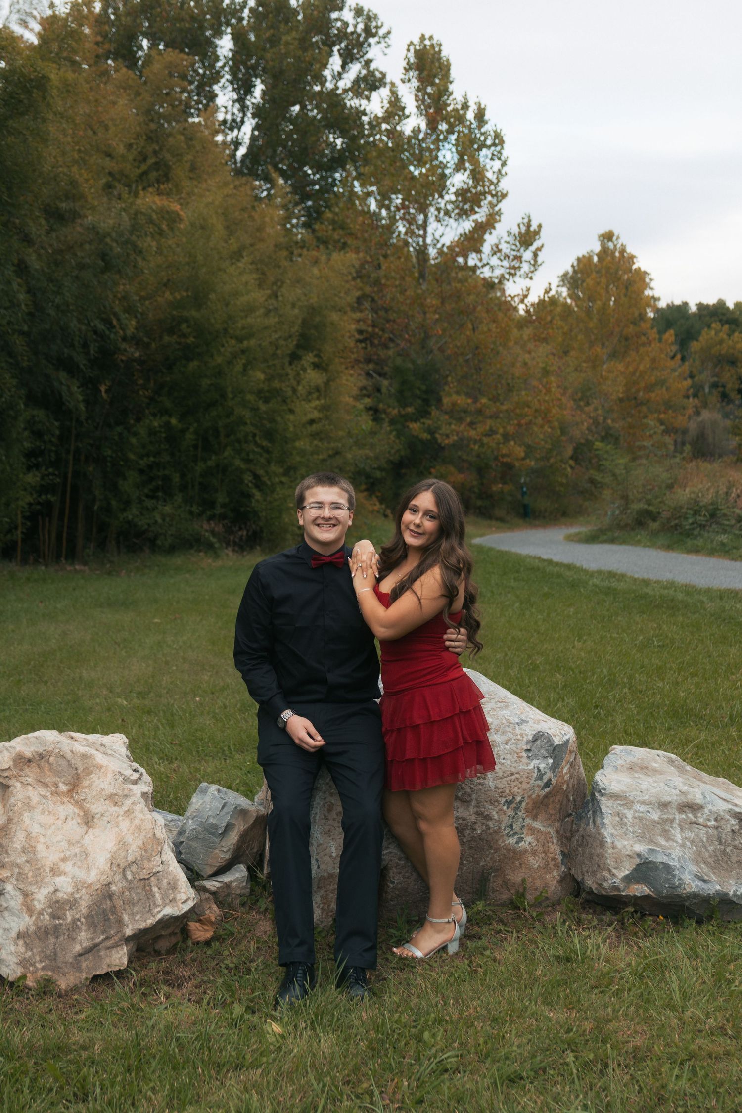 A young couple poses outdoors near large rocks with autumn trees, the girl in a red dress embracing a boy in all black.