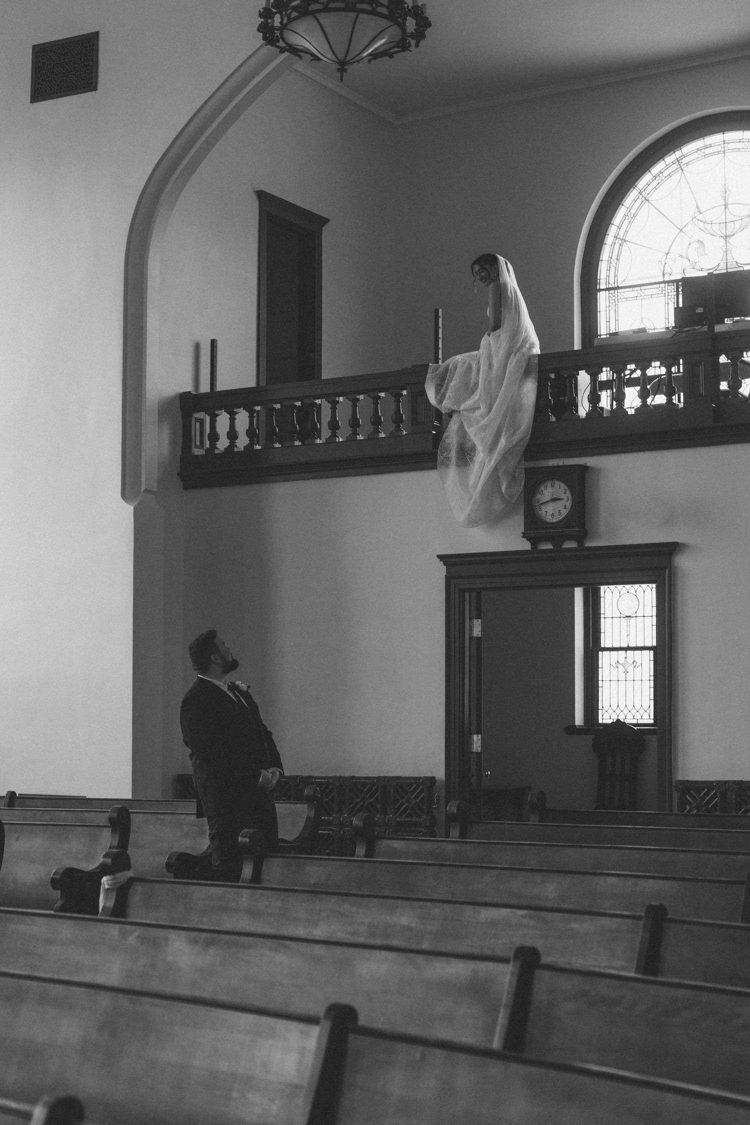 Black and white photo of a groom kneeling in church pews as bride stands on balcony above in wedding veil.