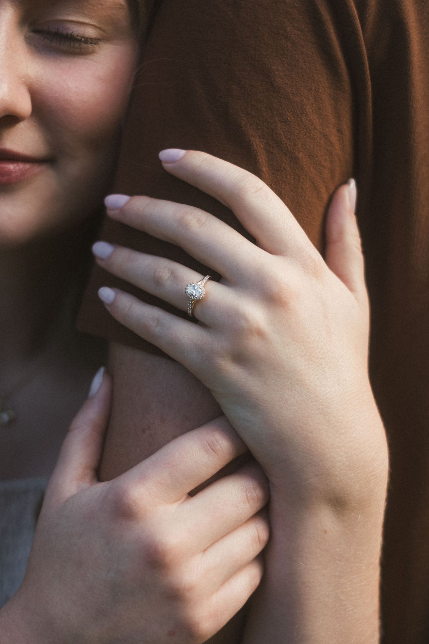 Close-up of a woman's hand wearing a delicate diamond engagement ring while embracing a partner in a brown shirt.