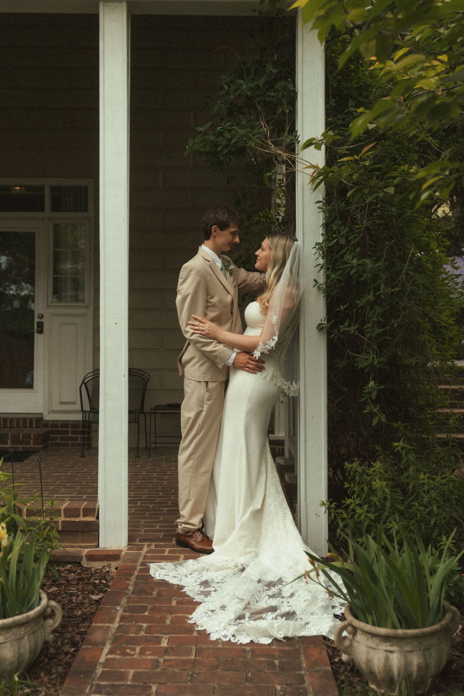 Bride in white lace gown and veil embraces groom in tan suit on brick porch surrounded by lush greenery.
