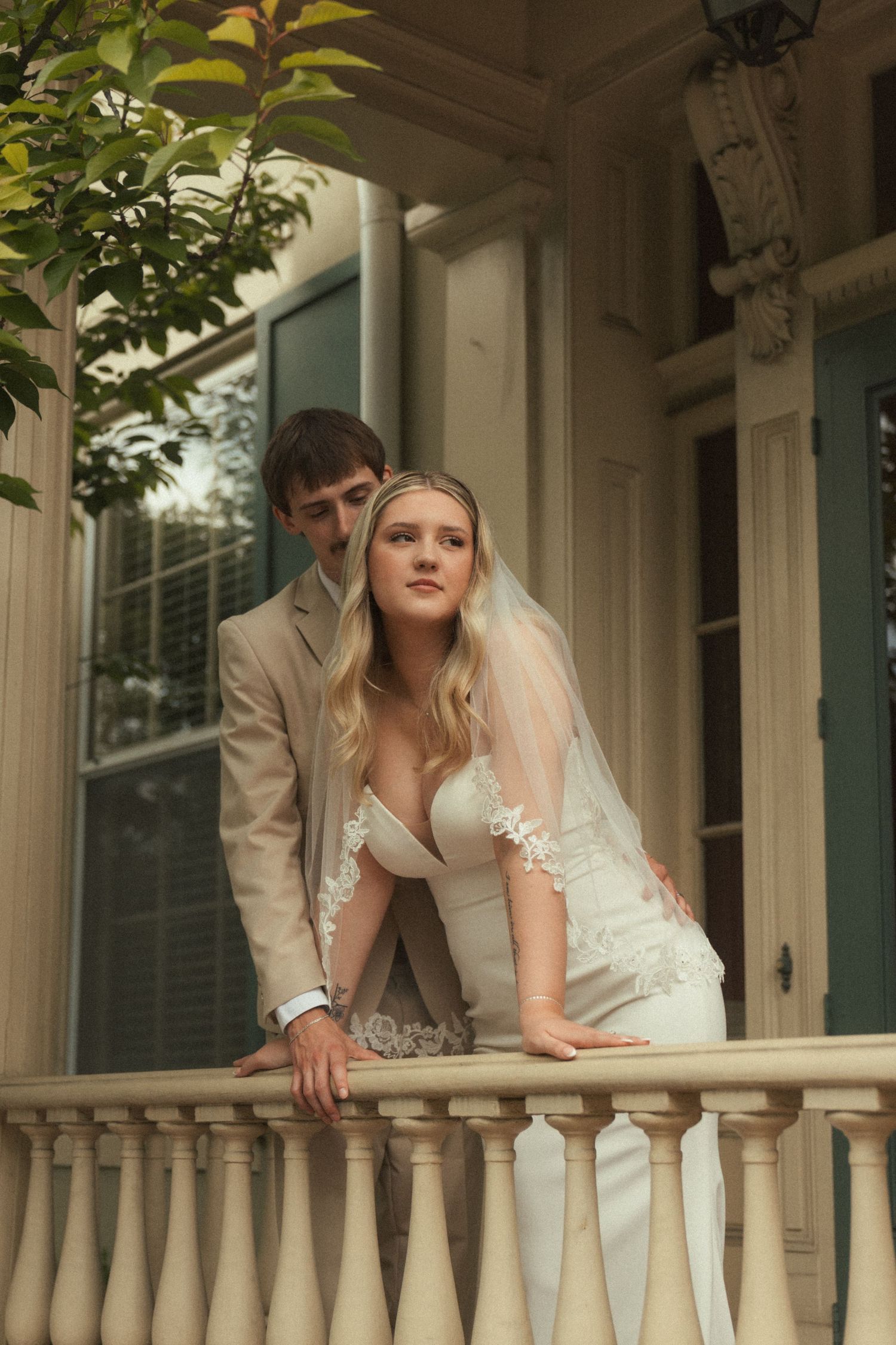 A bride in a lace veil and groom in a tan suit pose on a white balcony of an elegant Southern mansion.