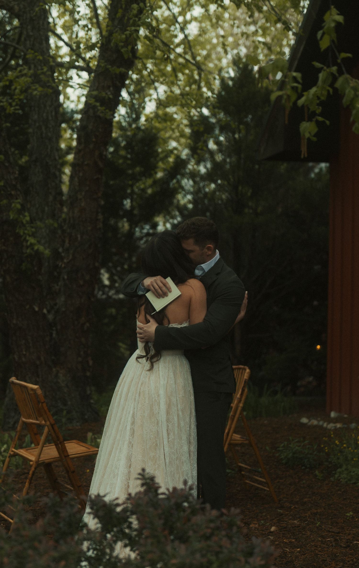 Bride and groom share a heartfelt embrace during an outdoor wedding ceremony surrounded by trees at dusk.