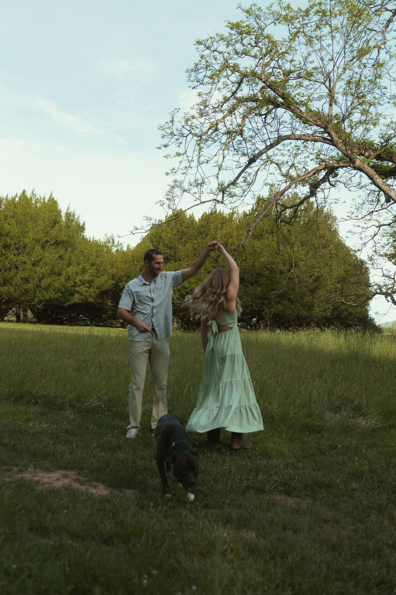 Couple dancing outdoors on green grass with a dog nearby, surrounded by lush trees under a blue sky.