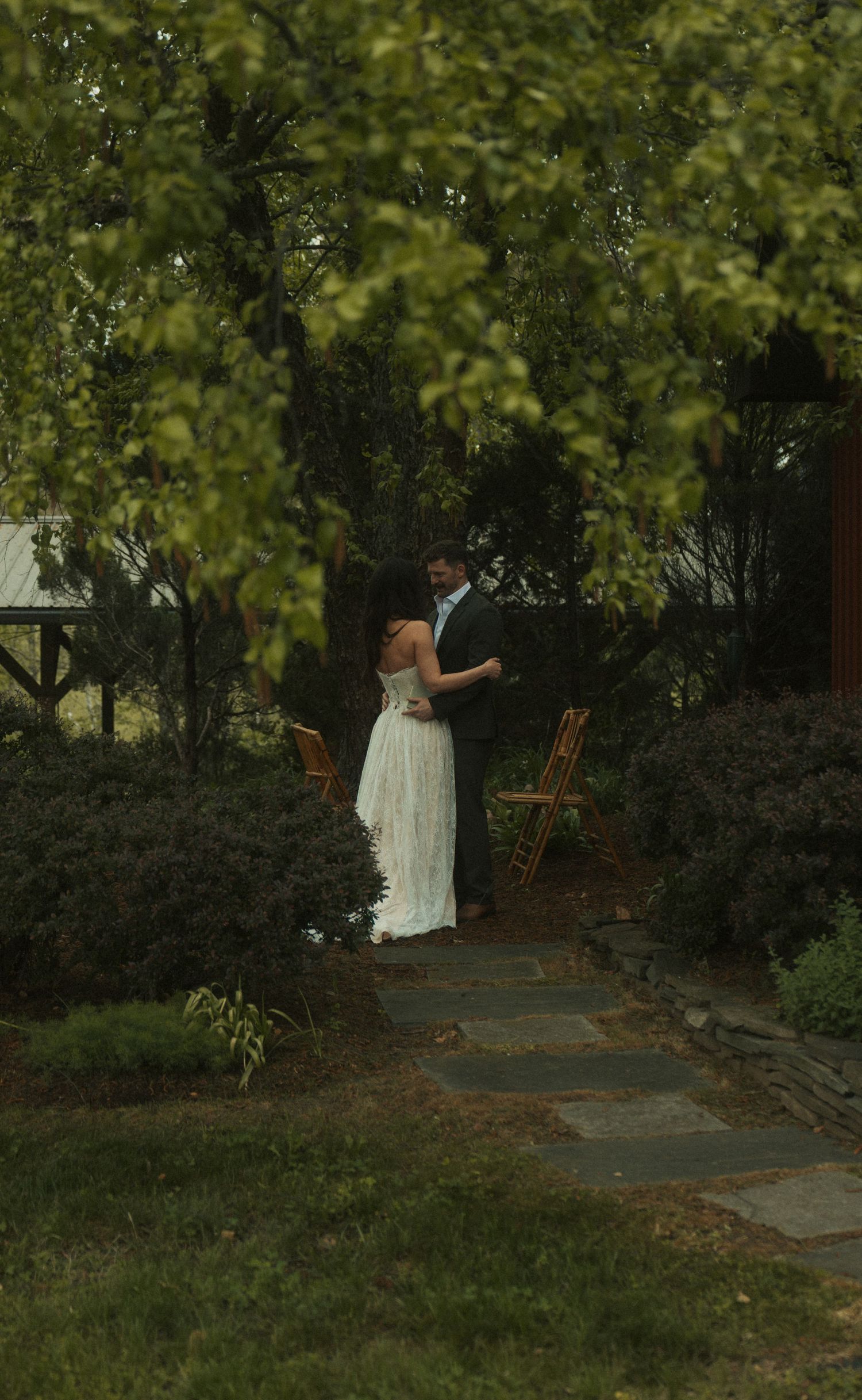 A couple in wedding attire shares a romantic dance on a stone path surrounded by lush green garden trees.