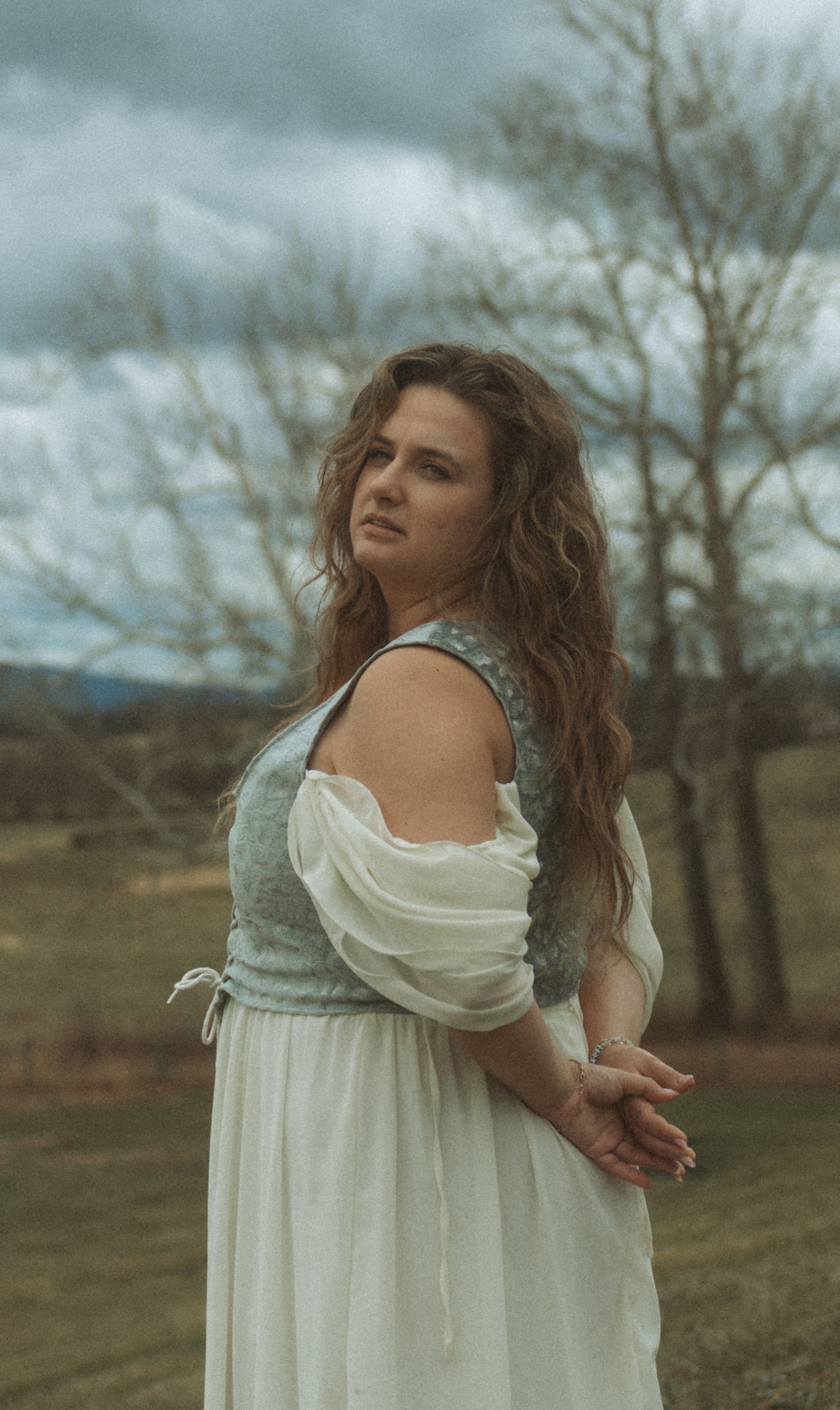 A person in a flowing white dress twirls gracefully in a moody outdoor setting with bare trees in the background.