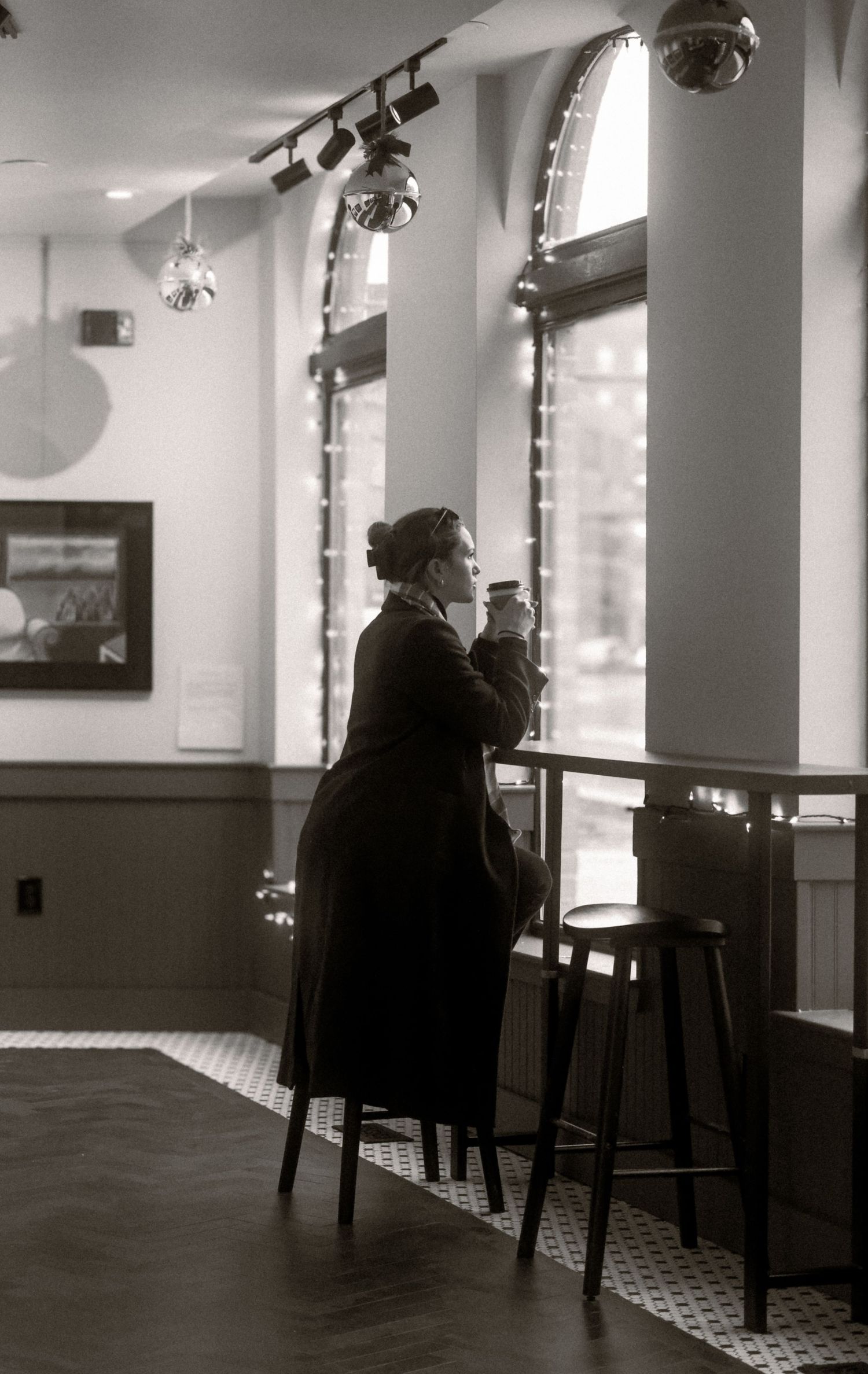 A silhouetted figure in a long coat drinks coffee while gazing out arched windows in a cafe with track lighting.