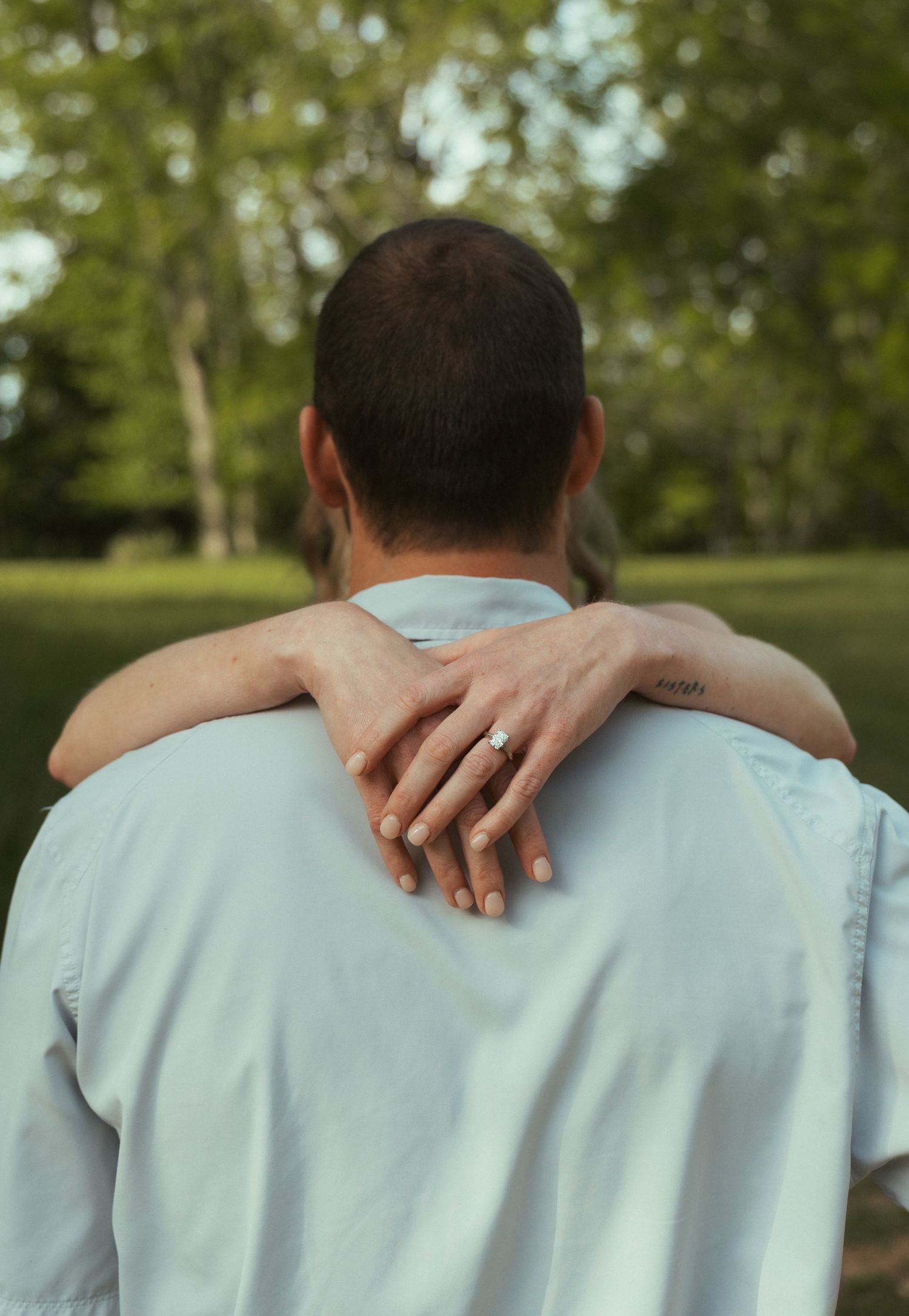 A couple embraces outdoors, showcasing an engagement ring on the woman's hand draped over her partner's shoulder.