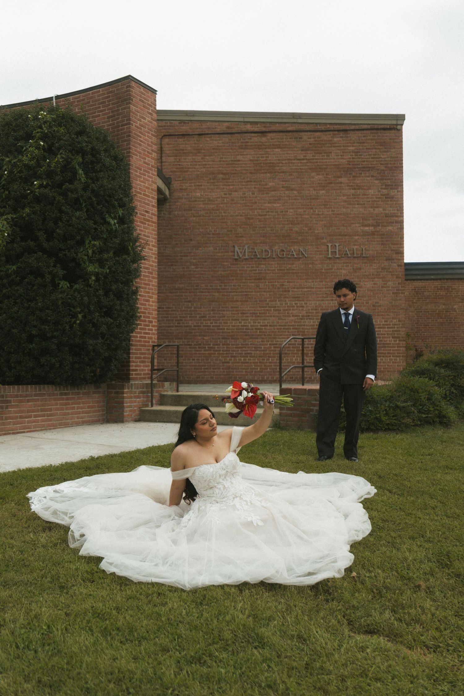 Bride in white gown sits on grass holding red bouquet while groom stands behind near brick Madatan Hall building.