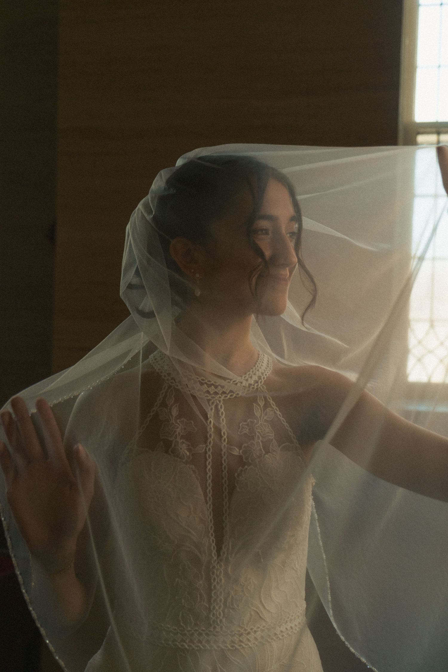 Smiling bride in lace wedding dress and flowing veil posed by window in warm natural light.