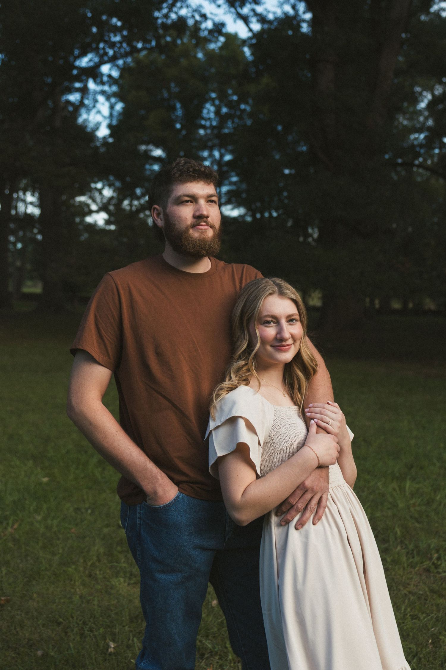 A couple poses outdoors in a park, man in brown tee hugs woman in white dress, trees in background.