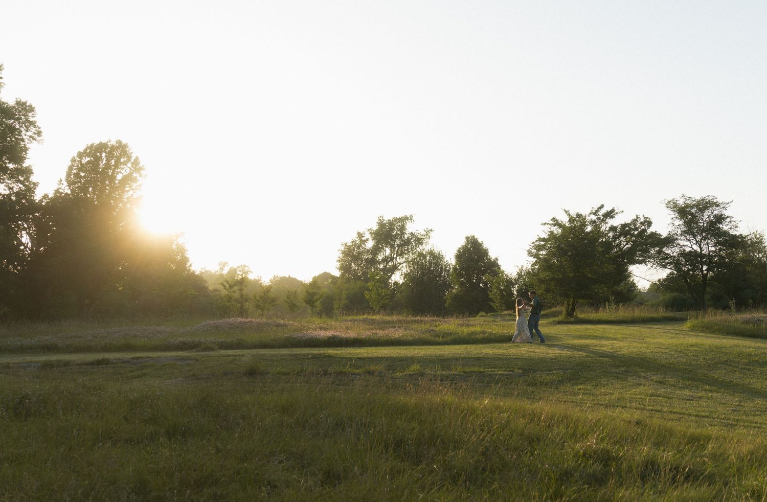 A couple stands embracing in a sunlit green meadow surrounded by trees during golden hour sunset.